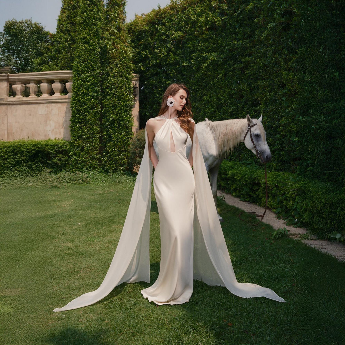 May include: A woman wearing a white halter-neck gown with a flowing cape, standing in a garden setting with a white horse in the background.