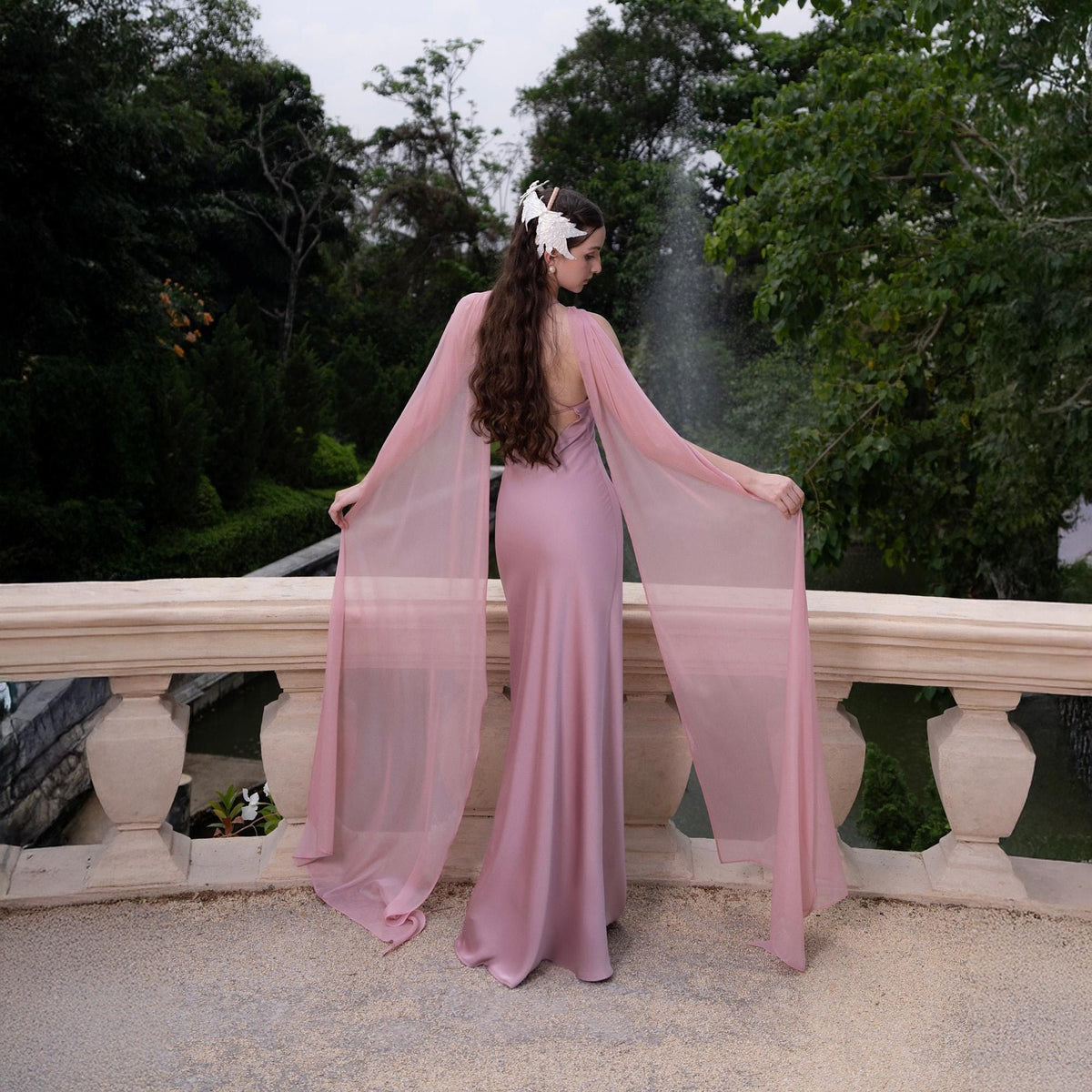 May include: A woman wearing a long, pink satin dress with a sheer pink cape. The dress has a low back and the cape is draped over her shoulders. She is standing on a stone balcony with a fountain in the background.