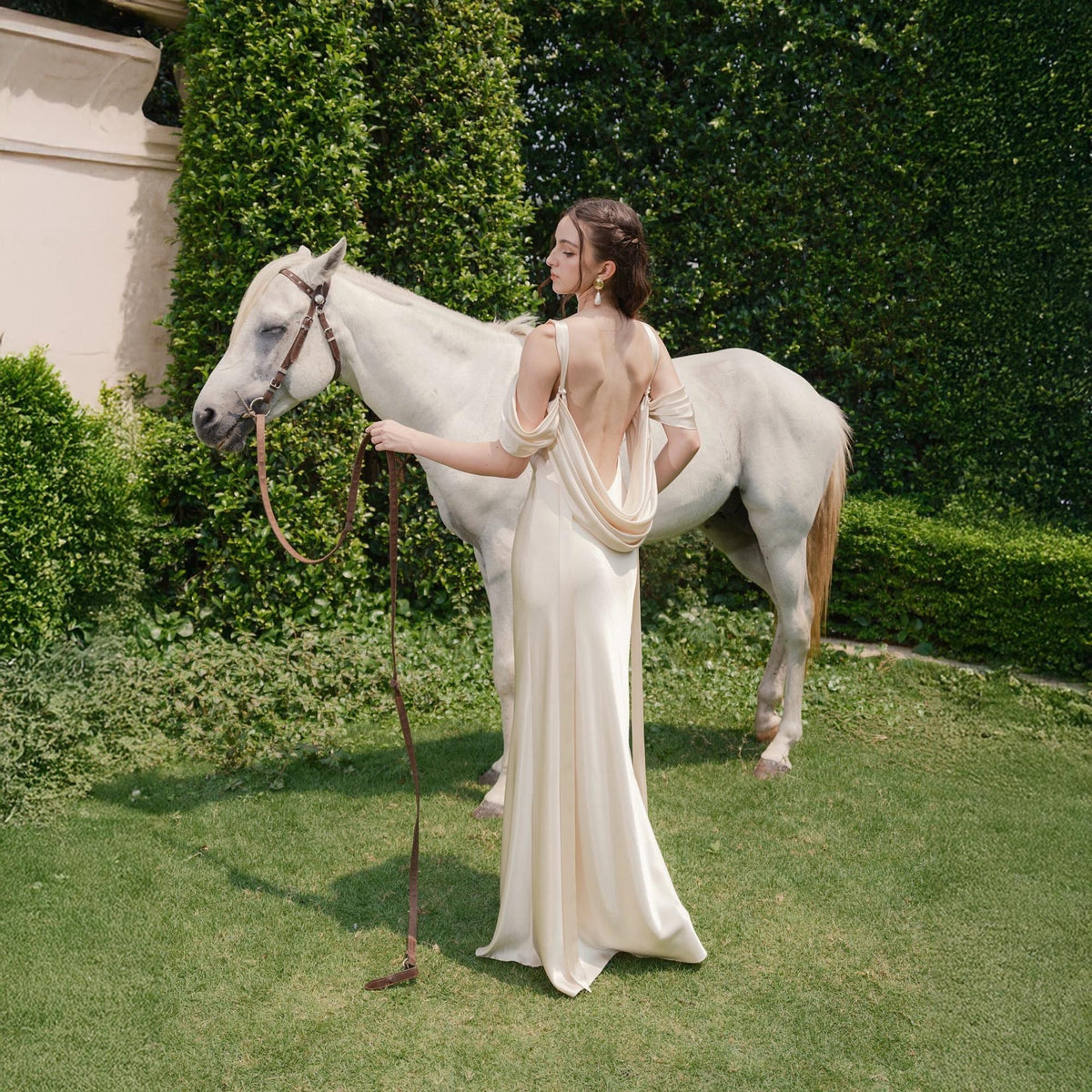 May include: A woman in a cream-colored, open-back gown stands next to a white horse on a grassy lawn. The dress features draped sleeves and a flowing skirt. The horse has a brown bridle and reins. Green hedges and trees form the background.