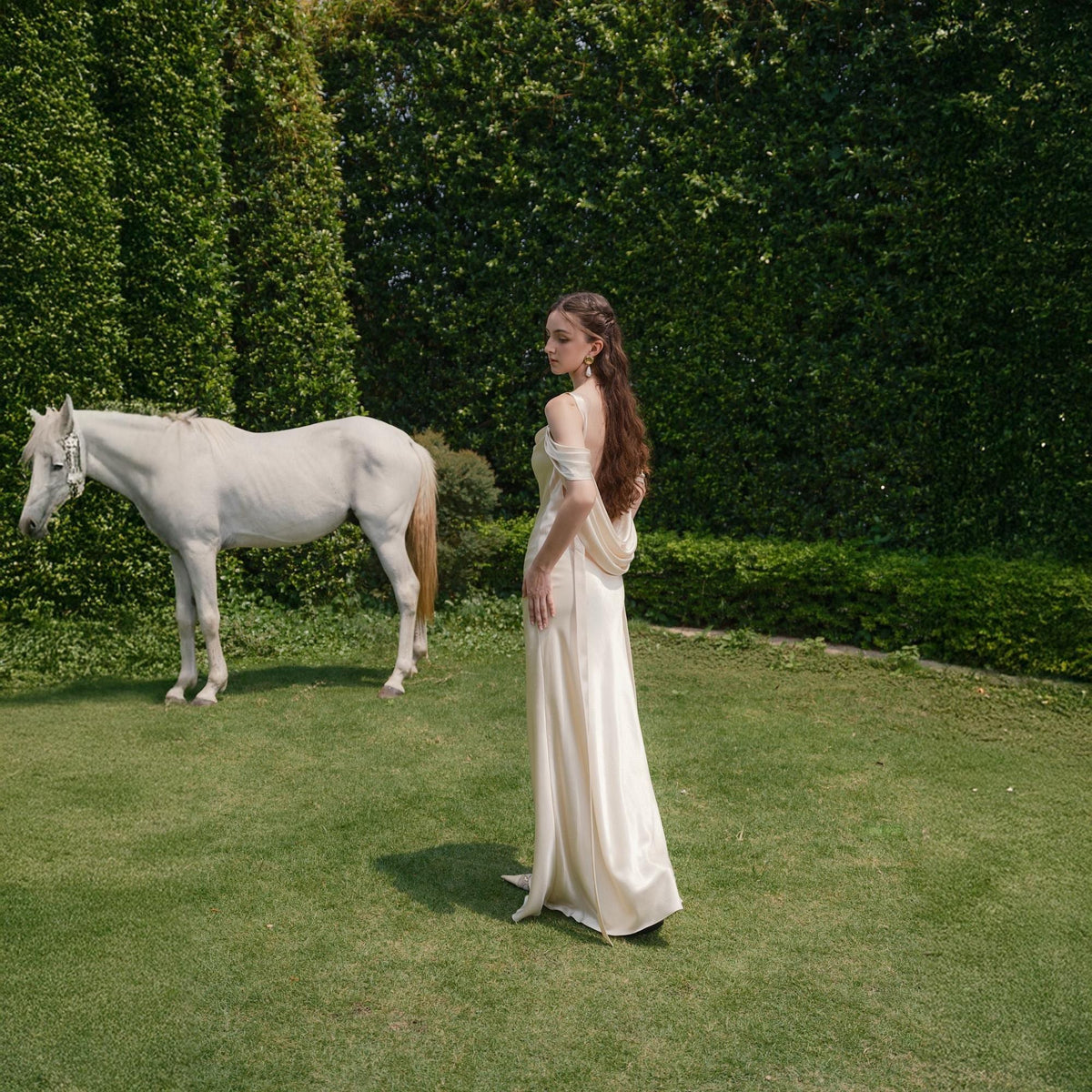 May include: A woman in a cream-colored, off-the-shoulder gown stands on a grassy lawn next to a white horse. The horse has a tan mane and tail. A tall, green hedge forms the background.