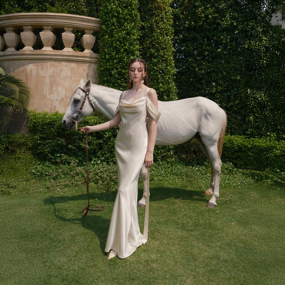 May include: A cream-colored, floor-length dress with a cowl neckline and spaghetti straps. The dress is worn by a person standing next to a white horse. The scene is set outdoors with green grass and foliage.