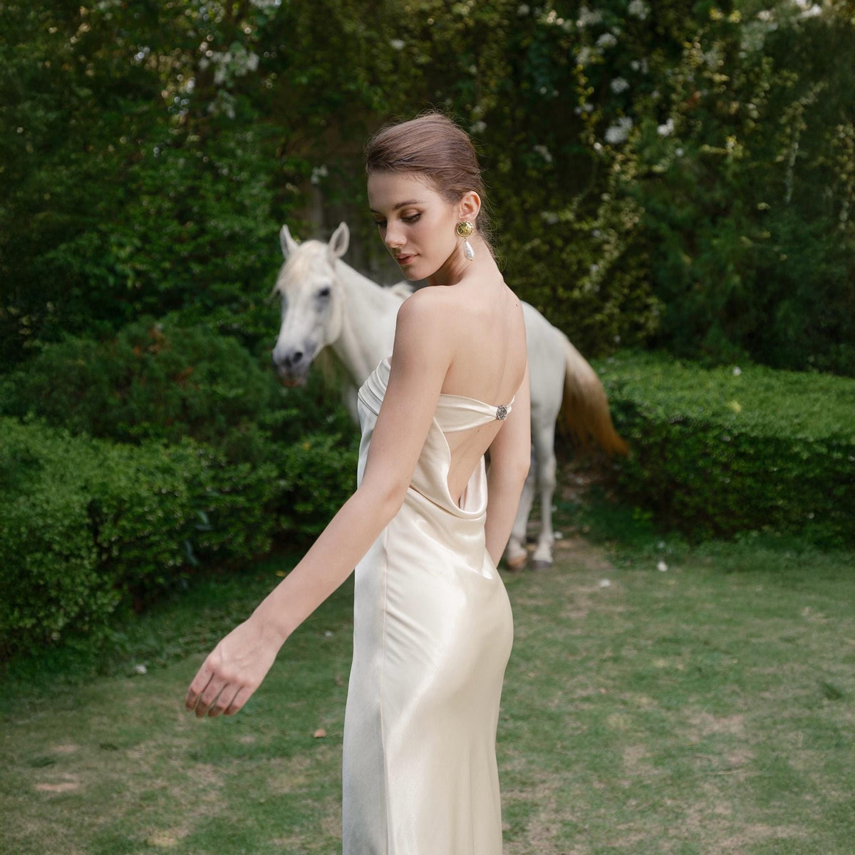May include: A woman in a cream-colored satin gown with a low back poses outdoors. A white horse stands in the background on a grassy lawn, surrounded by green bushes and trees. The dress has a draped neckline and a subtle sheen.