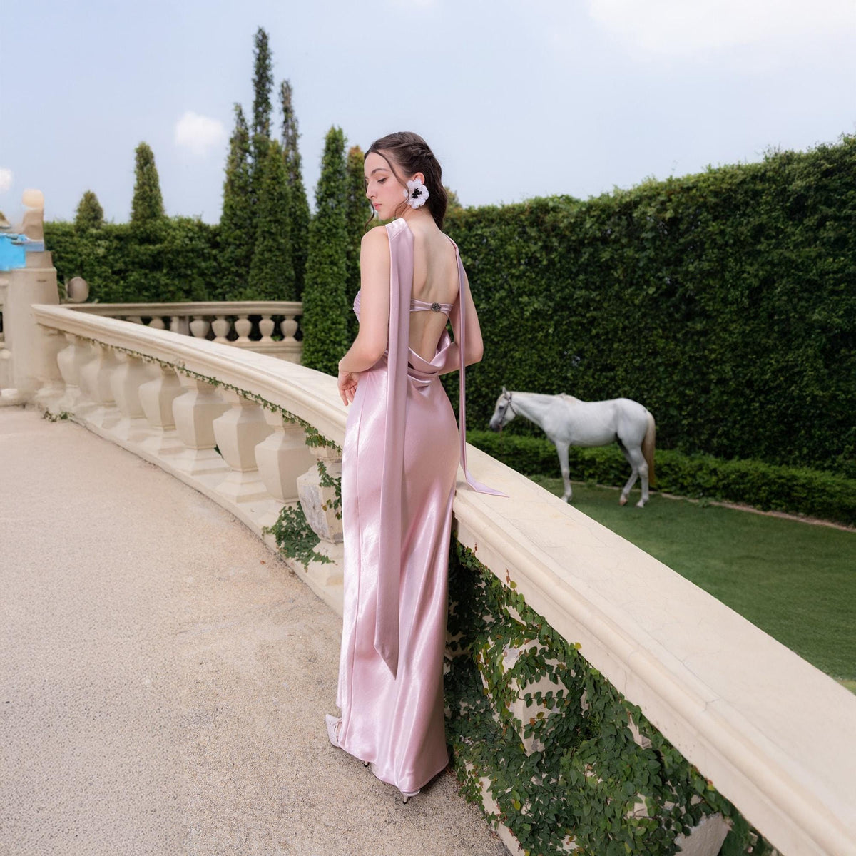 May include: A light pink satin gown with a flowing design. The dress features a low back and long, draping fabric that extends from the shoulders. The model is standing on a stone walkway with a white horse in the background.
