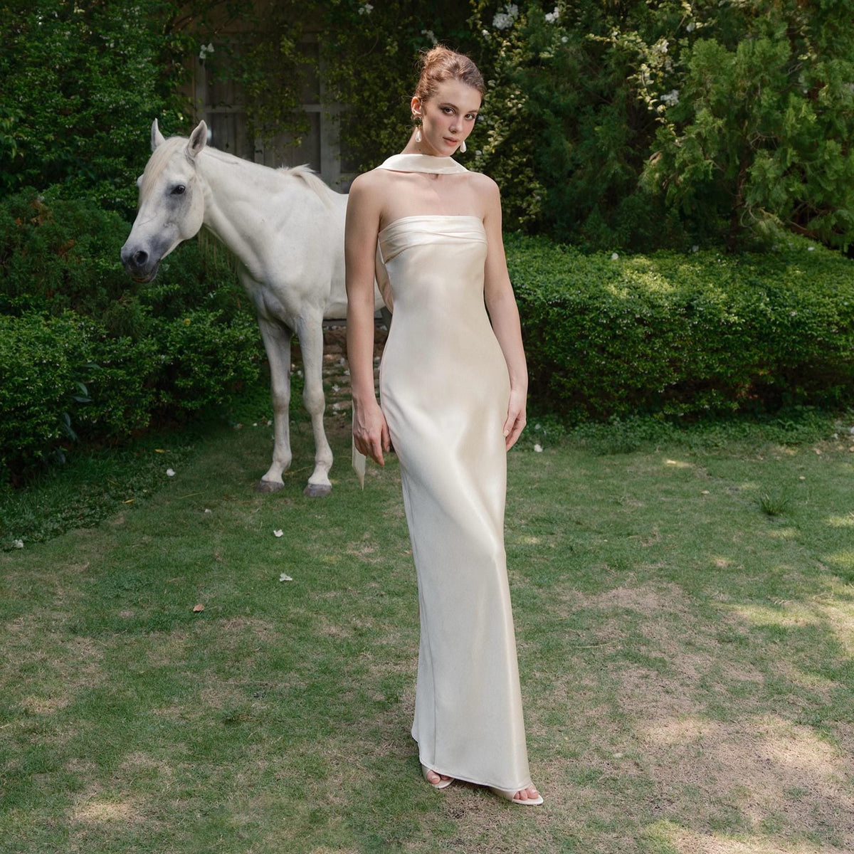 May include: A strapless, floor-length ivory gown with a flowing silhouette. The dress features a draped bodice and a matching scarf. A white horse stands in the background on a grassy lawn, surrounded by green bushes.