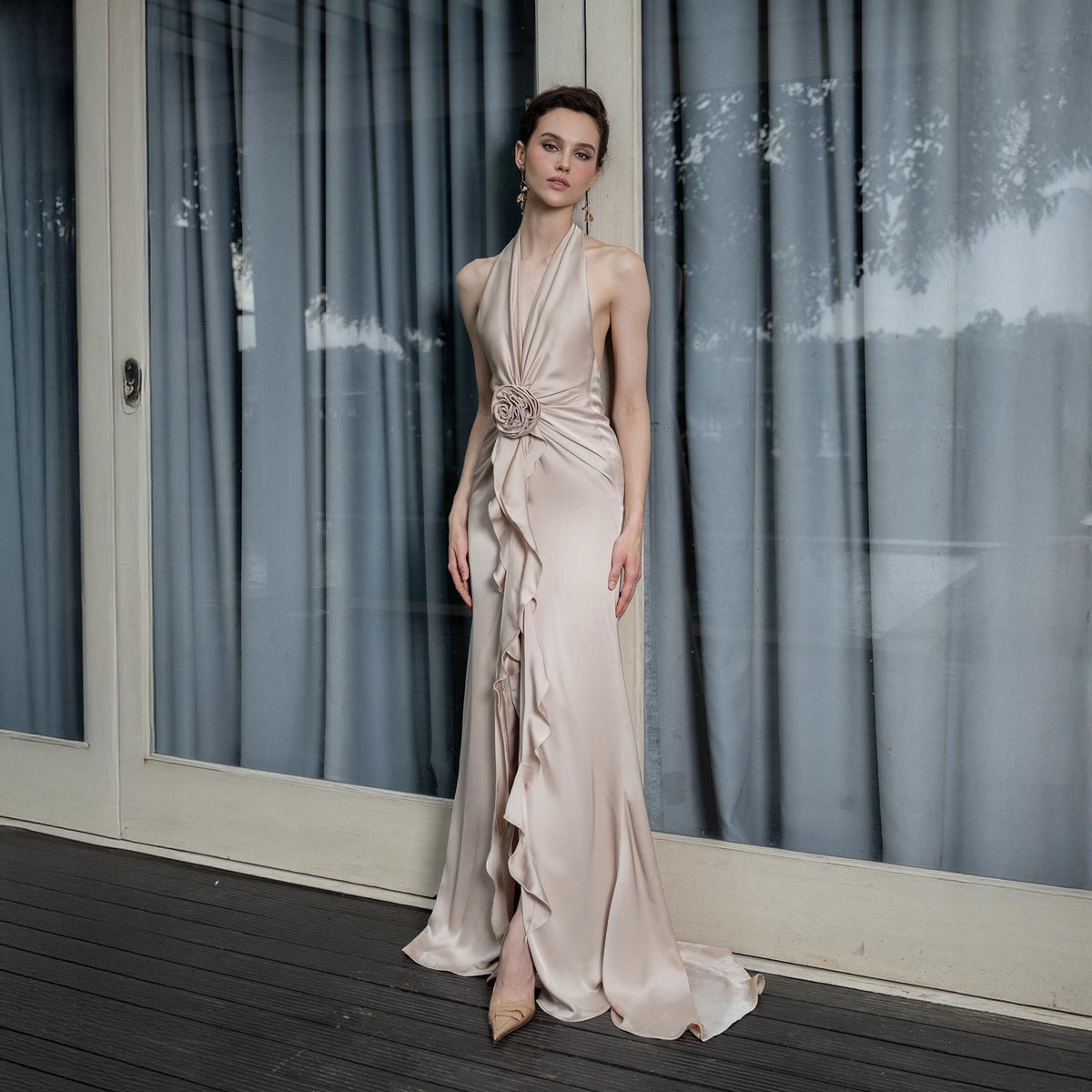 May include: A champagne-colored, floor-length gown with a halter neckline and a flowing skirt. The dress features a decorative floral embellishment at the waist and ruffled detailing down the front. The dress is made of a silky material.