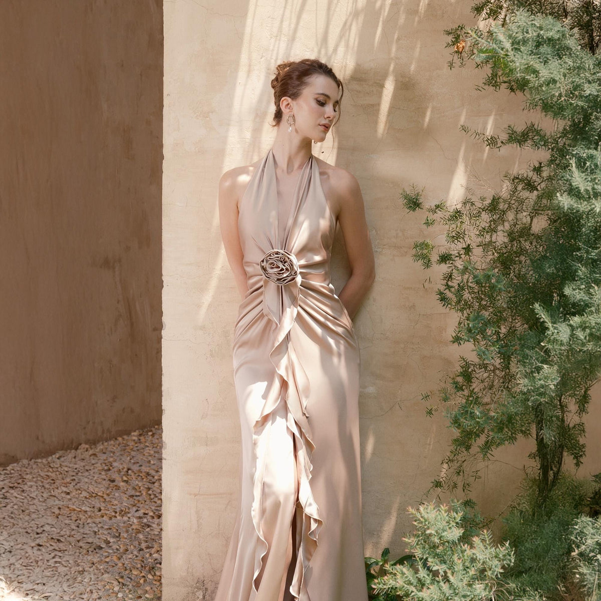 May include: A champagne-colored halter-neck gown with a flowing silhouette. The dress features a decorative embellishment at the waist and cascading ruffles down the front. The elegant design is suitable for formal events.