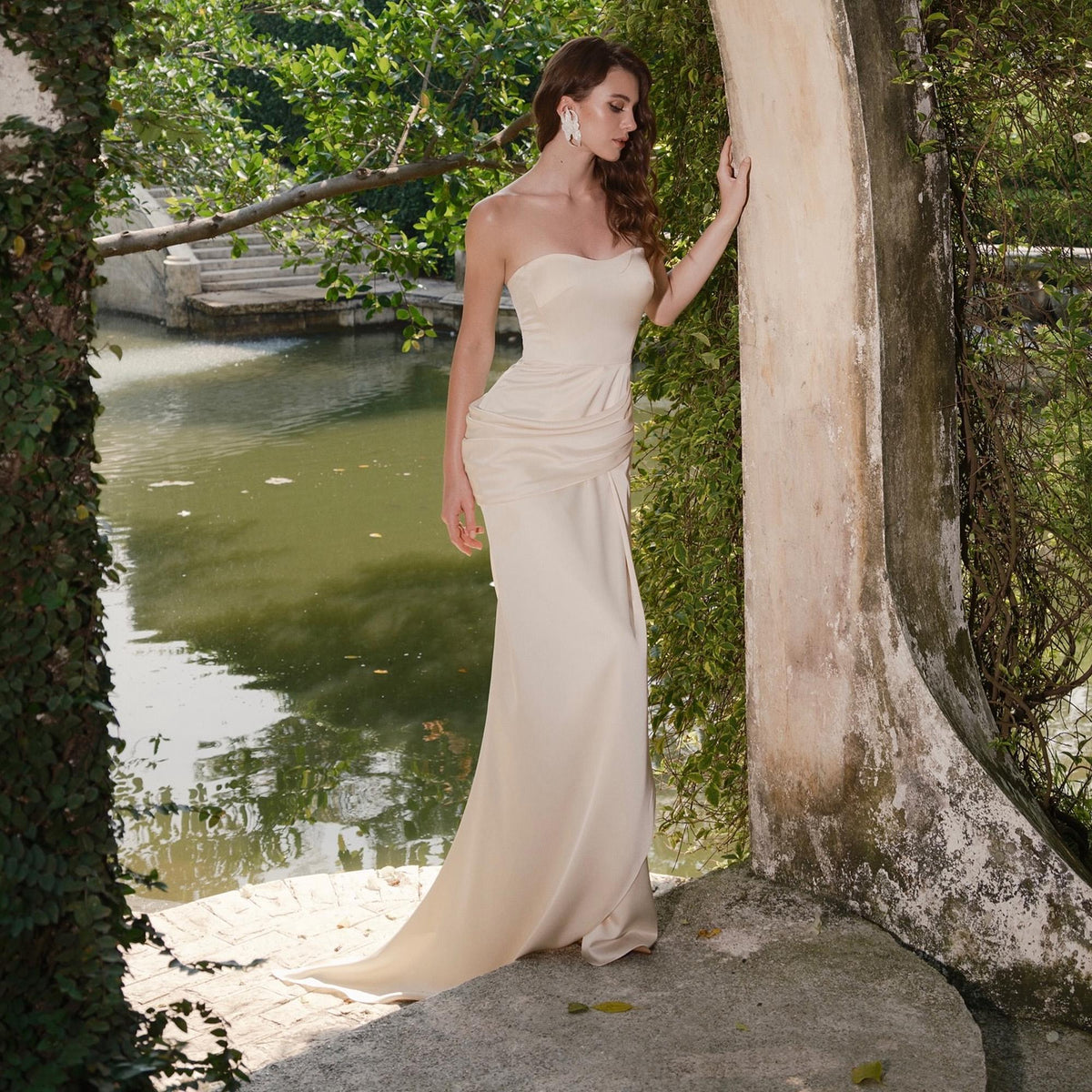 May include: A strapless, floor-length ivory gown with a draped bodice and a flowing skirt. The dress is worn outdoors near a body of water, with greenery in the background. The gown's elegant design is suitable for formal events.