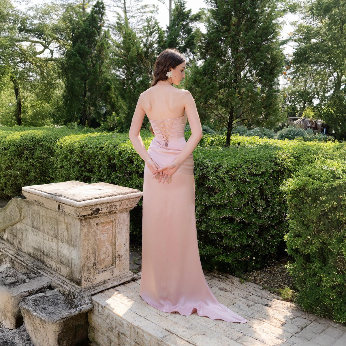 May include: A strapless, pale pink formal gown with a fitted bodice and a flowing skirt. The back of the dress features a lace-up detail. The dress is worn outdoors, with a garden and stone structure in the background.