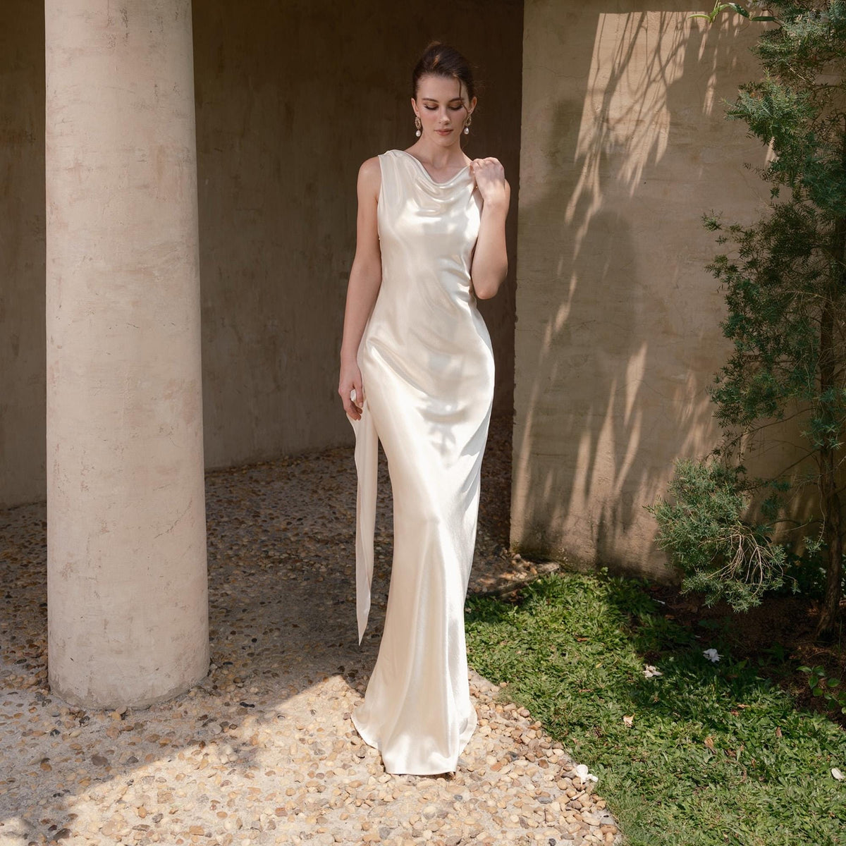 May include: A sleeveless, ivory-colored satin gown with a cowl neckline and a flowing, floor-length skirt. The dress features a side slit and a long, flowing sash. The model is standing in an outdoor setting with a stone floor and a concrete pillar.