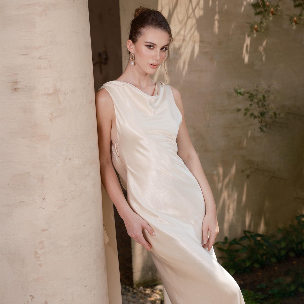 May include: A sleeveless, ivory-colored dress with a cowl neckline. The dress has a flowing, silky texture and drapes elegantly. The model is leaning against a beige column in a sunlit outdoor setting. The dress appears to be floor-length.