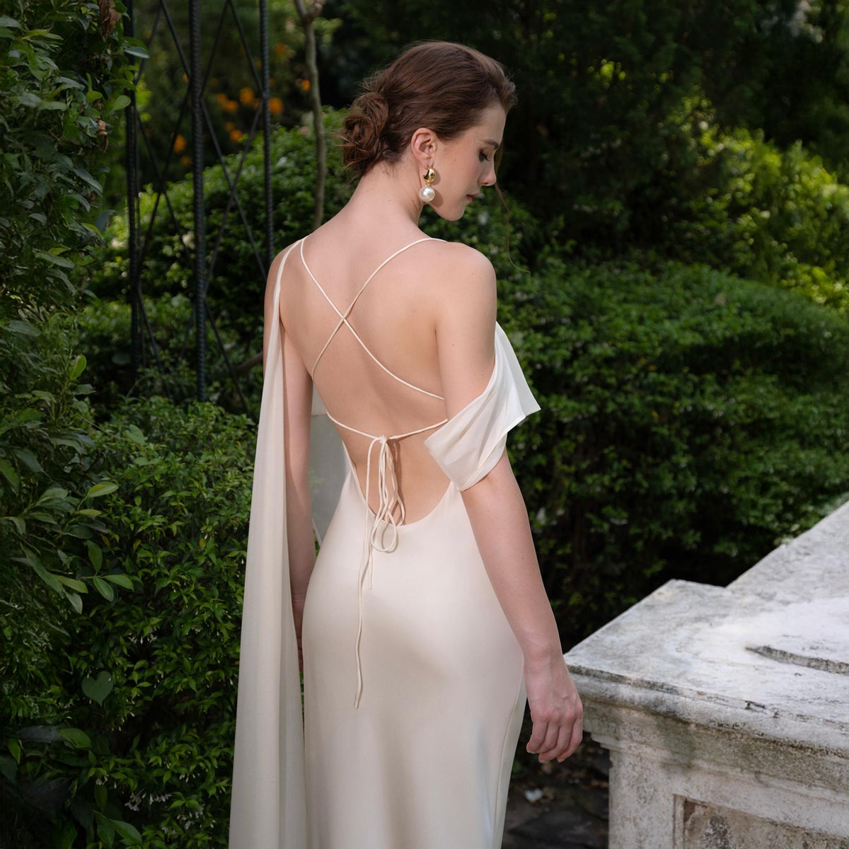 May include: A cream-colored, backless gown with spaghetti straps that crisscross the back. The dress features a flowing cape-like detail on one side. The model is wearing gold earrings and is standing in a garden setting.