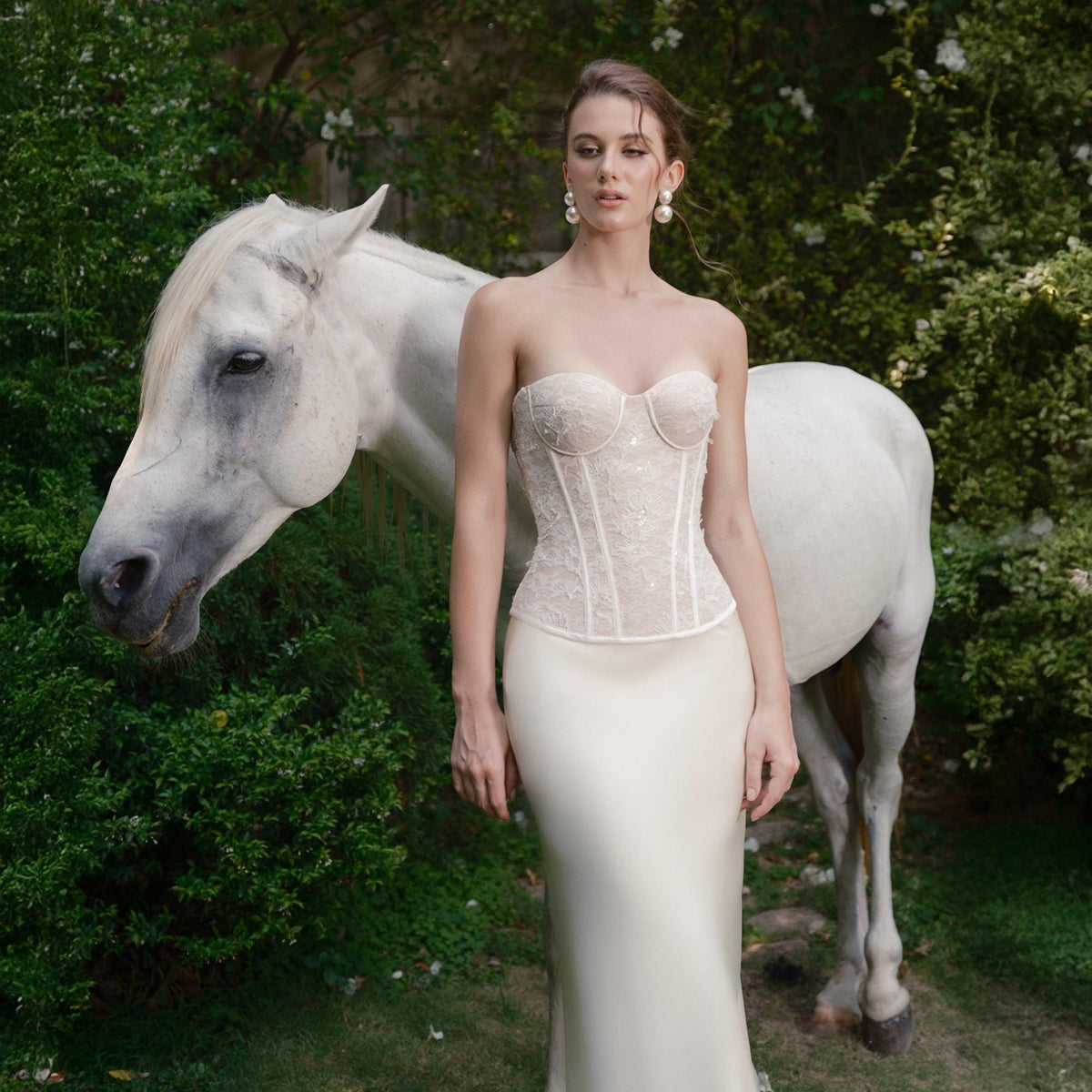May include: A strapless ivory wedding gown with a fitted bodice and a flowing skirt. The bodice is embellished with delicate lace and sequins. A white horse stands beside the person wearing the gown, with green foliage in the background.