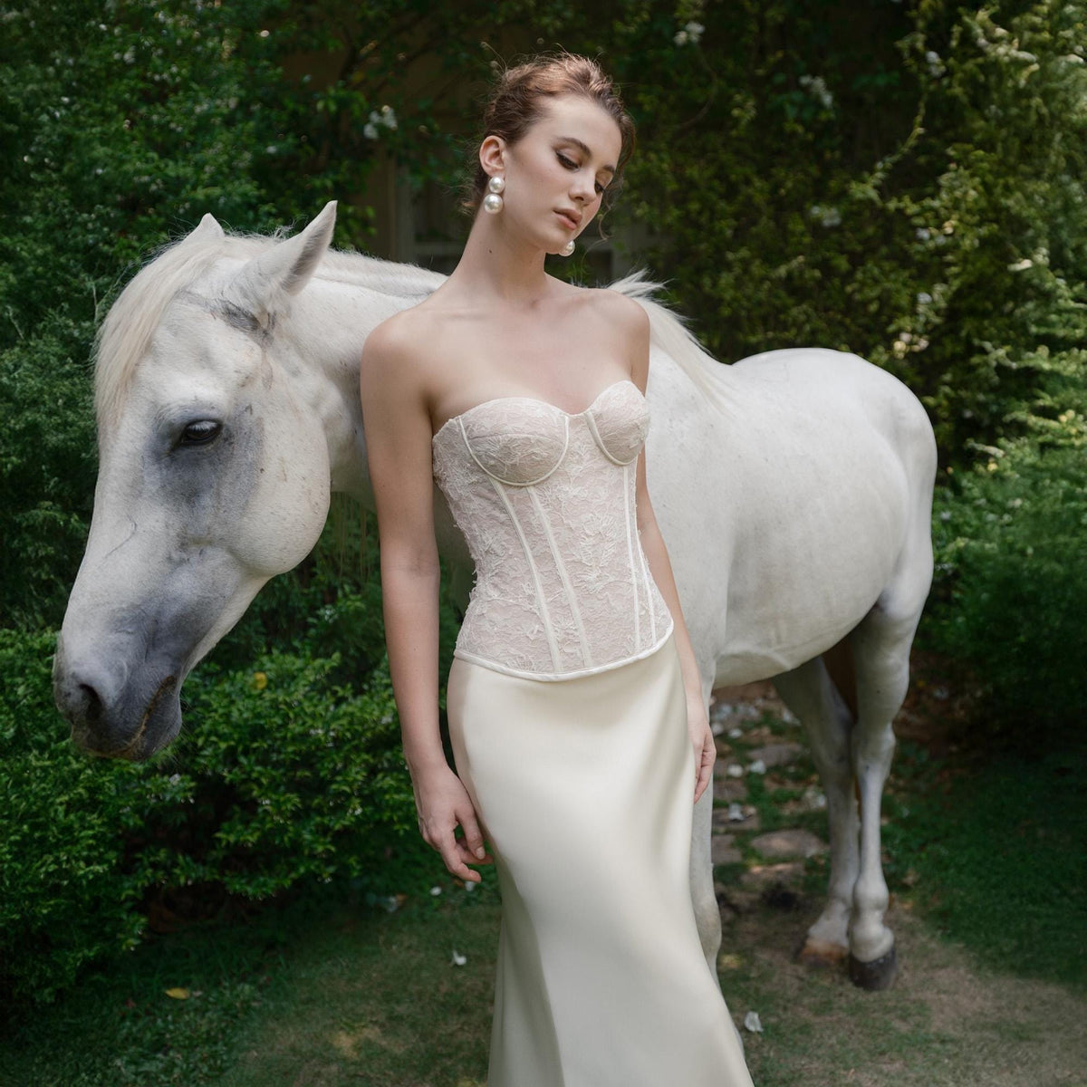 May include: A strapless ivory corset with delicate lace detailing, paired with a flowing satin skirt. A white horse stands nearby in a lush green garden setting. The model is wearing pearl earrings.