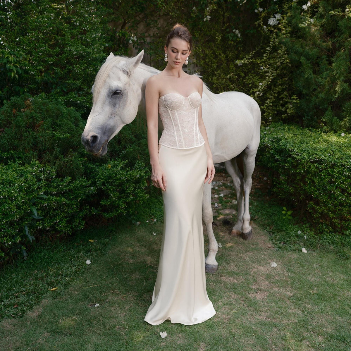 May include: A strapless ivory wedding gown with a lace bodice and a flowing satin skirt. The model is standing next to a white horse in a garden setting. The dress is elegant and suitable for a formal event.
