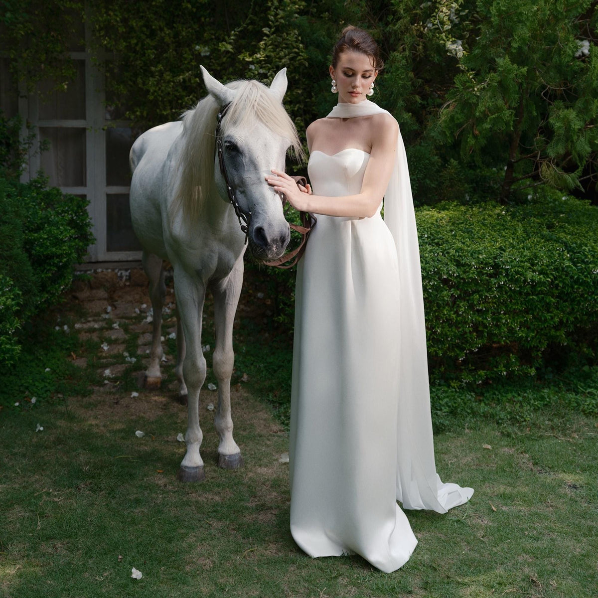 May include: A strapless white gown with a flowing cape, worn by a person standing next to a white horse. The dress has a simple, elegant design. The scene is set outdoors with green foliage in the background, creating a serene and natural setting.