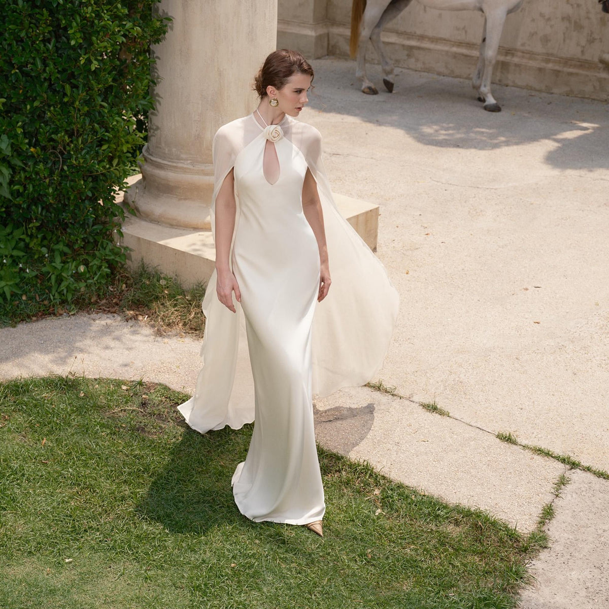 May include: A white satin wedding gown with a keyhole neckline and a flowing cape. The dress features a floral embellishment at the neckline. The bride is standing on a green lawn next to a stone walkway.