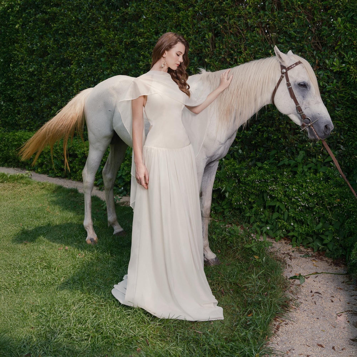 May include: A cream-colored, flowing gown with a high neckline and sheer sleeves. A white horse stands beside the gown. The scene is set outdoors with green foliage in the background. The horse has a brown bridle.