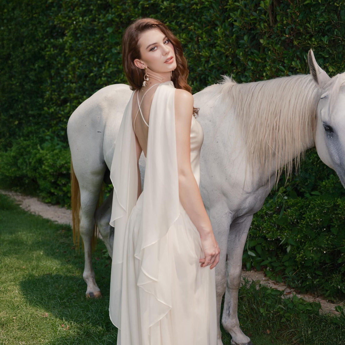 May include: A cream-colored, flowing gown with a draped back and layered skirt. The dress is worn by a person standing next to a white horse. The background features green foliage and grass.