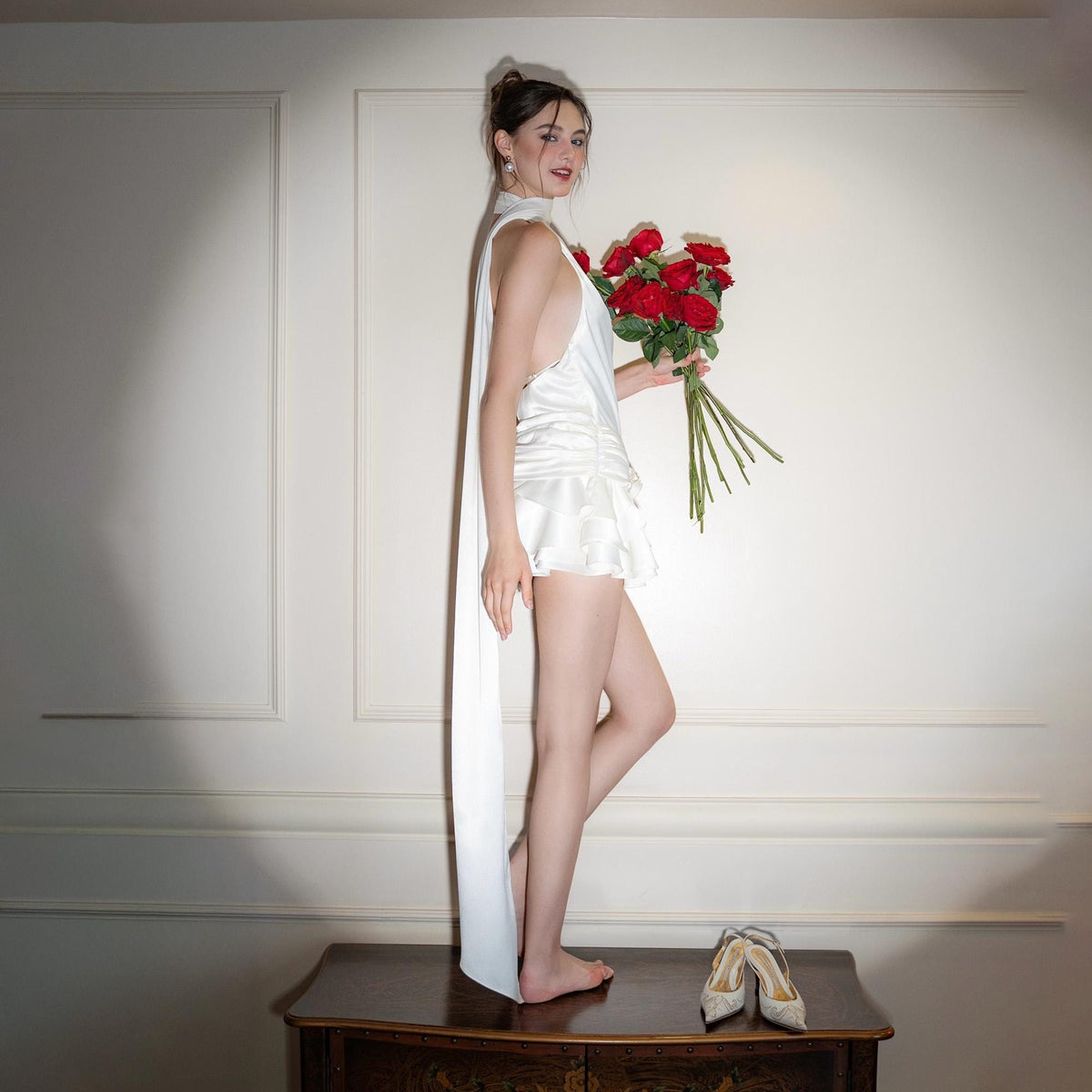 May include: A woman in a white satin dress with a halter neckline and a flowing sash, holding a bouquet of red roses. The dress has a ruffled skirt. A pair of silver heels are on a wooden surface.