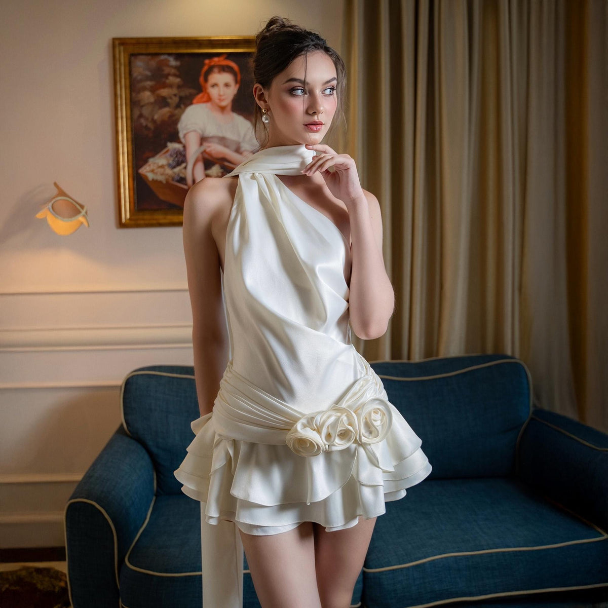 May include: A cream-colored satin dress with a halter neckline and a ruffled skirt. The dress features a decorative floral embellishment at the waist. The model is standing in front of a blue sofa and a painting.