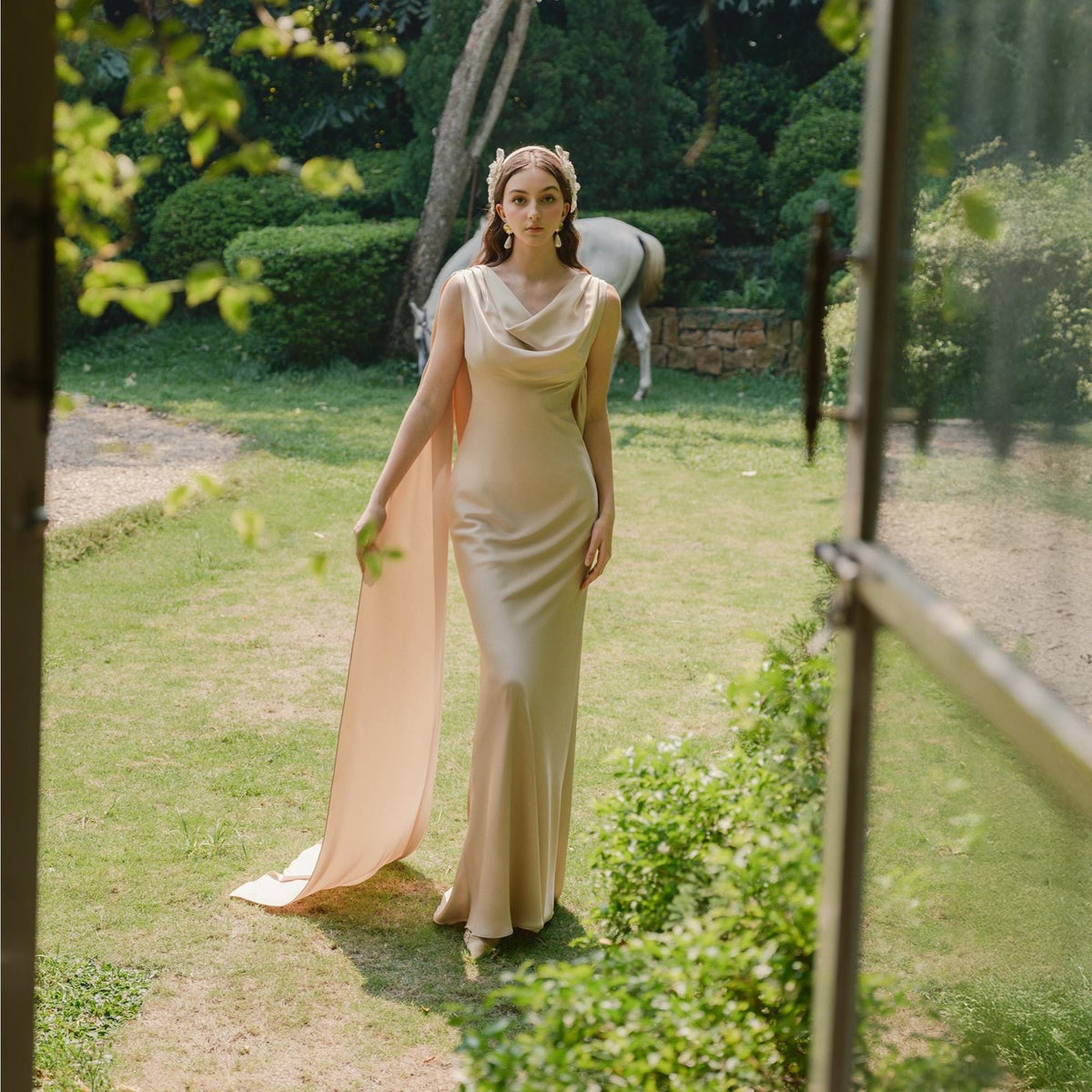 May include: A woman wearing a champagne-colored, floor-length gown with a cowl neckline and a flowing cape. The dress is made of a silky material. She is standing in a garden with a horse in the background. She is wearing a decorative headpiece.