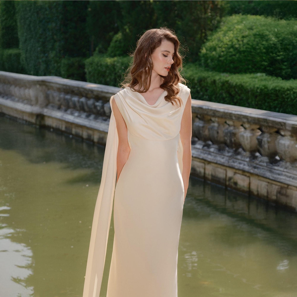 May include: A sleeveless, floor-length ivory gown with a draped cowl neckline and flowing cape-like sleeves. The dress is made of a smooth, elegant fabric. The background features a stone wall and a body of water.