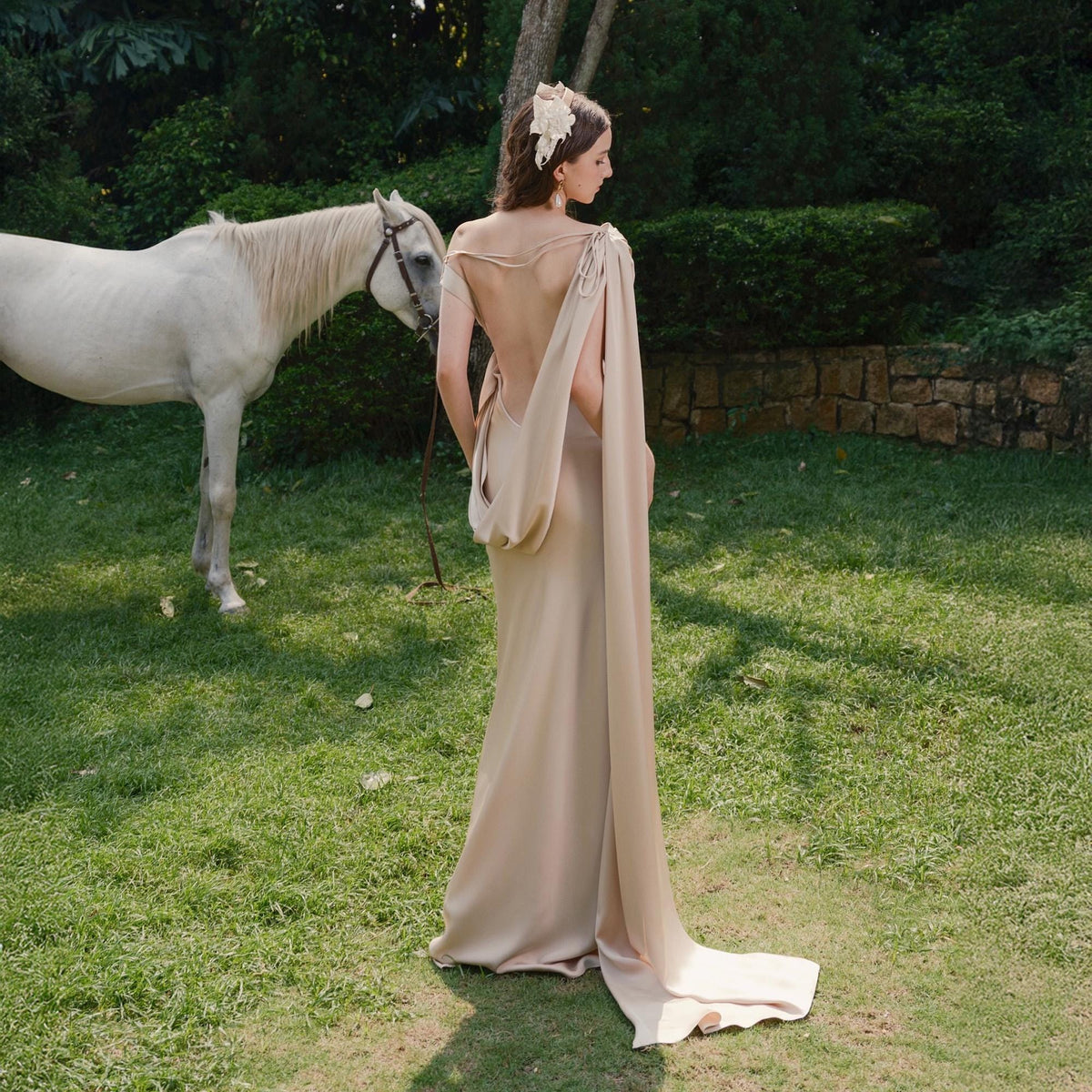 May include: A beige, floor-length gown with a draped back and flowing cape. A white horse stands in the background on a grassy lawn. The dress features a low back and a train. The model is wearing a floral hair accessory.