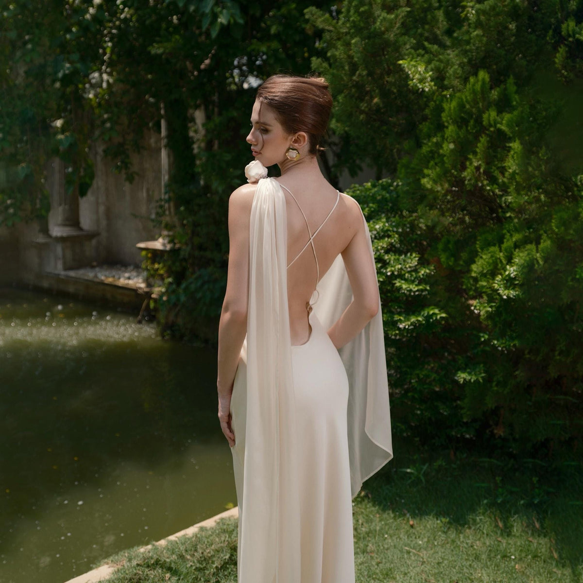 May include: A cream-colored, backless gown with a flowing cape. The dress features delicate spaghetti straps that crisscross the back. The model is standing near a pond with lush greenery in the background.
