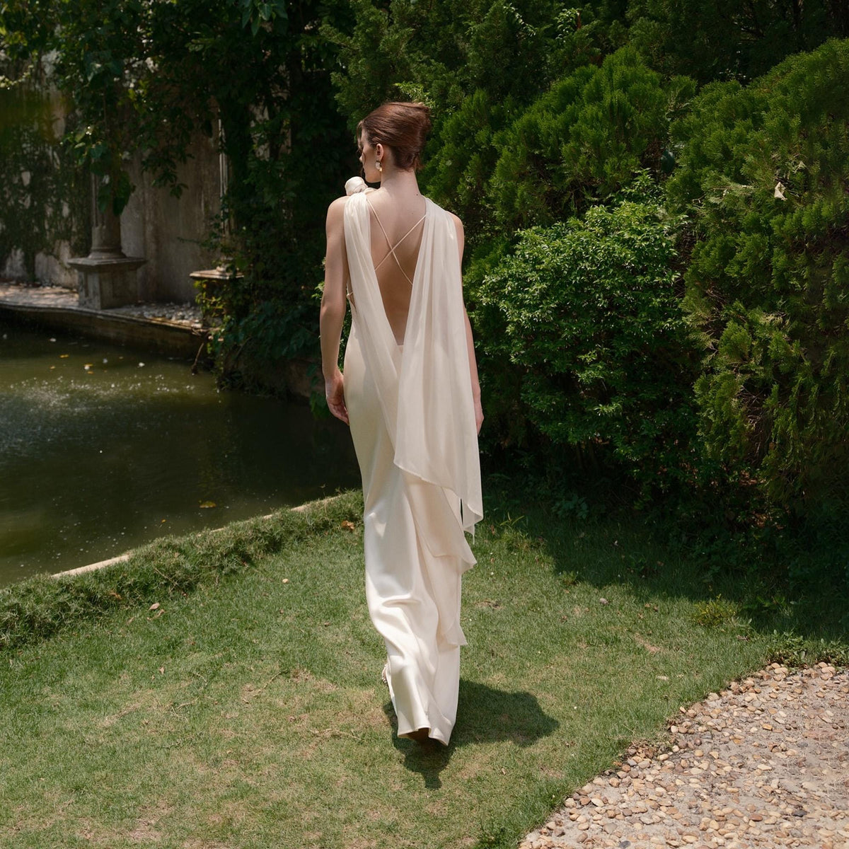 May include: A cream-colored, floor-length gown with a low back and draped fabric. The dress is worn outdoors, with a person walking on a grassy area near a pond and lush greenery.