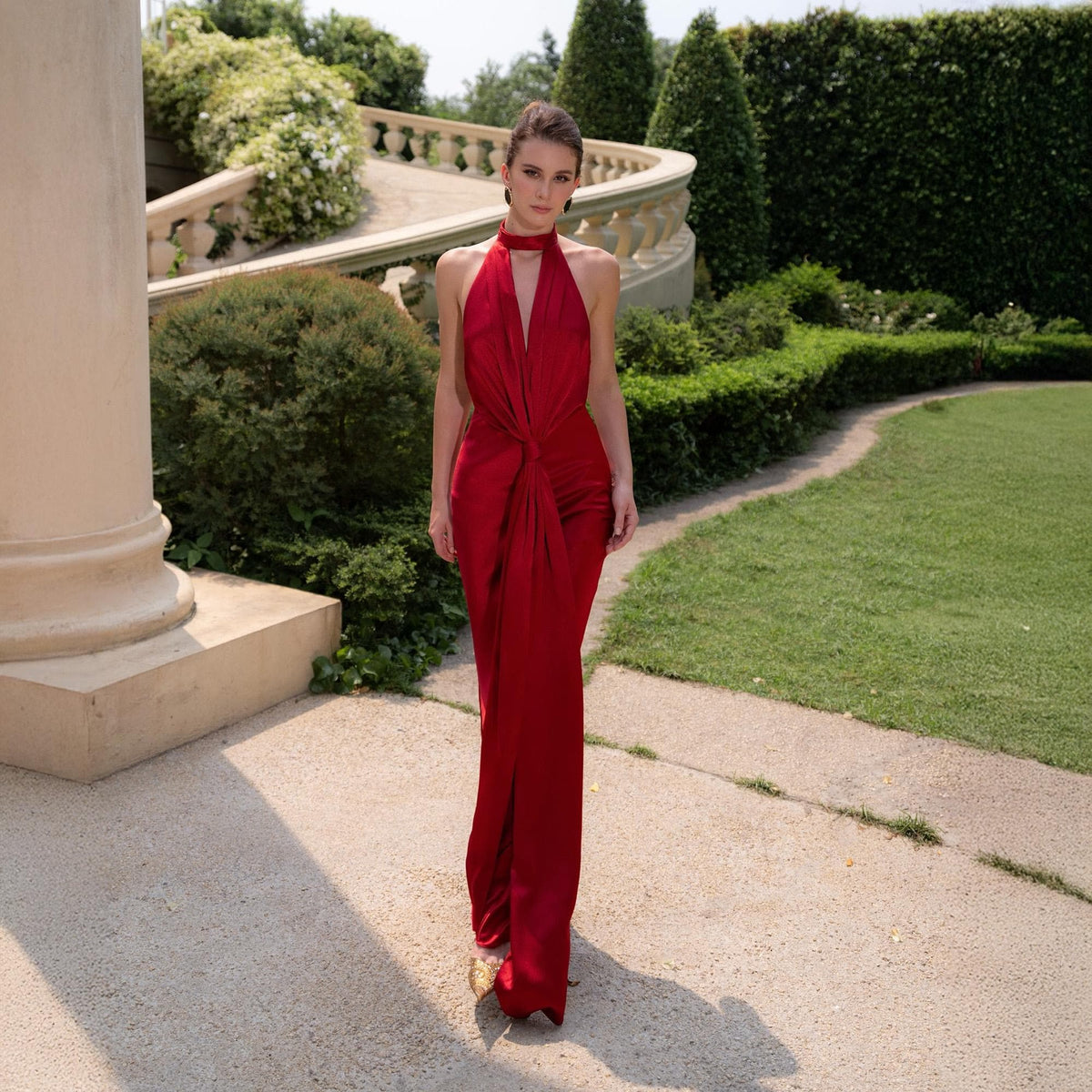 May include: A long, red halter dress with a knot detail at the waist. The dress has a flowing, floor-length skirt and a high neckline. The model is standing on a stone path in front of a garden.