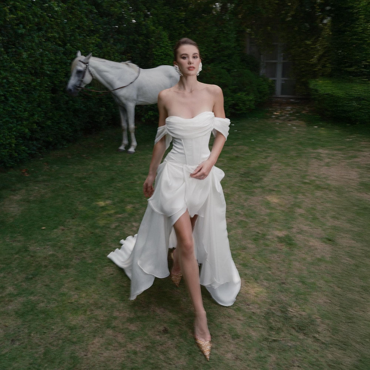 May include: A white off-the-shoulder wedding dress with a layered skirt and a high-low hemline. The dress is worn with gold-colored heels. A white horse stands in the background on a grassy lawn.