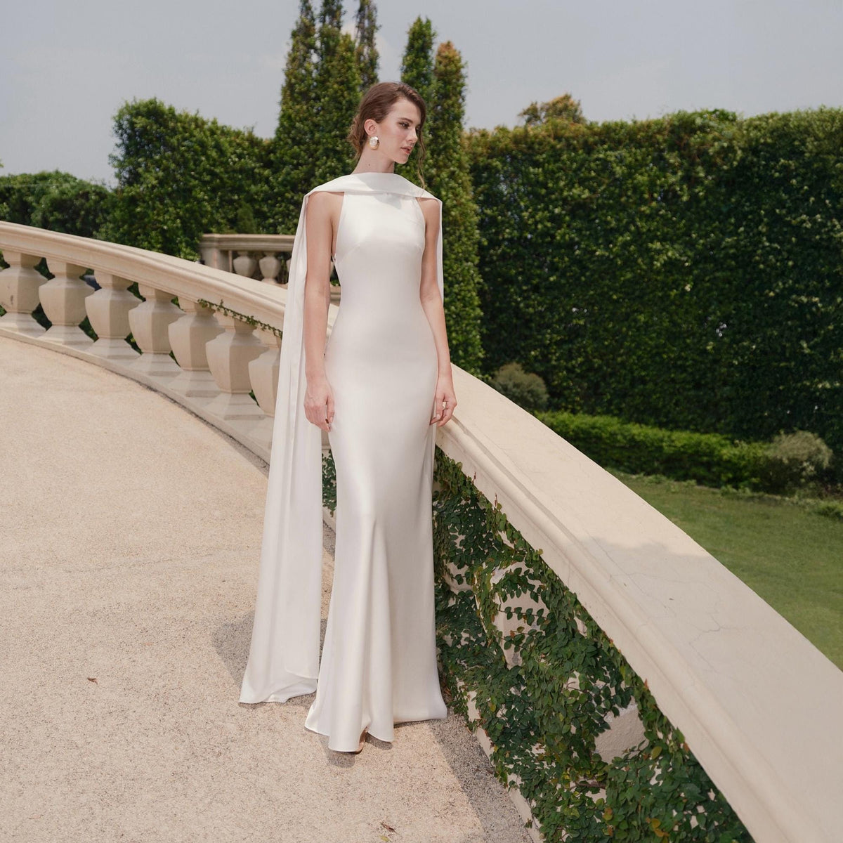 May include: A white, floor-length gown with a high neckline and a flowing cape. The dress has a fitted silhouette that flares out slightly at the hem. The model is standing on a stone walkway with a green hedge in the background.