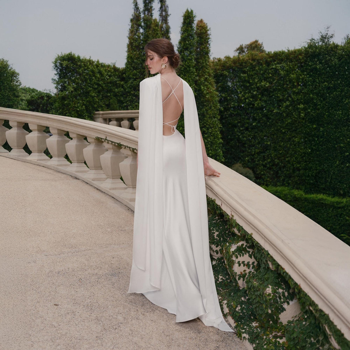 May include: A white satin gown with a flowing cape. The dress features a low back with crisscross straps. The model is standing on a stone walkway with a balustrade, with green foliage in the background.