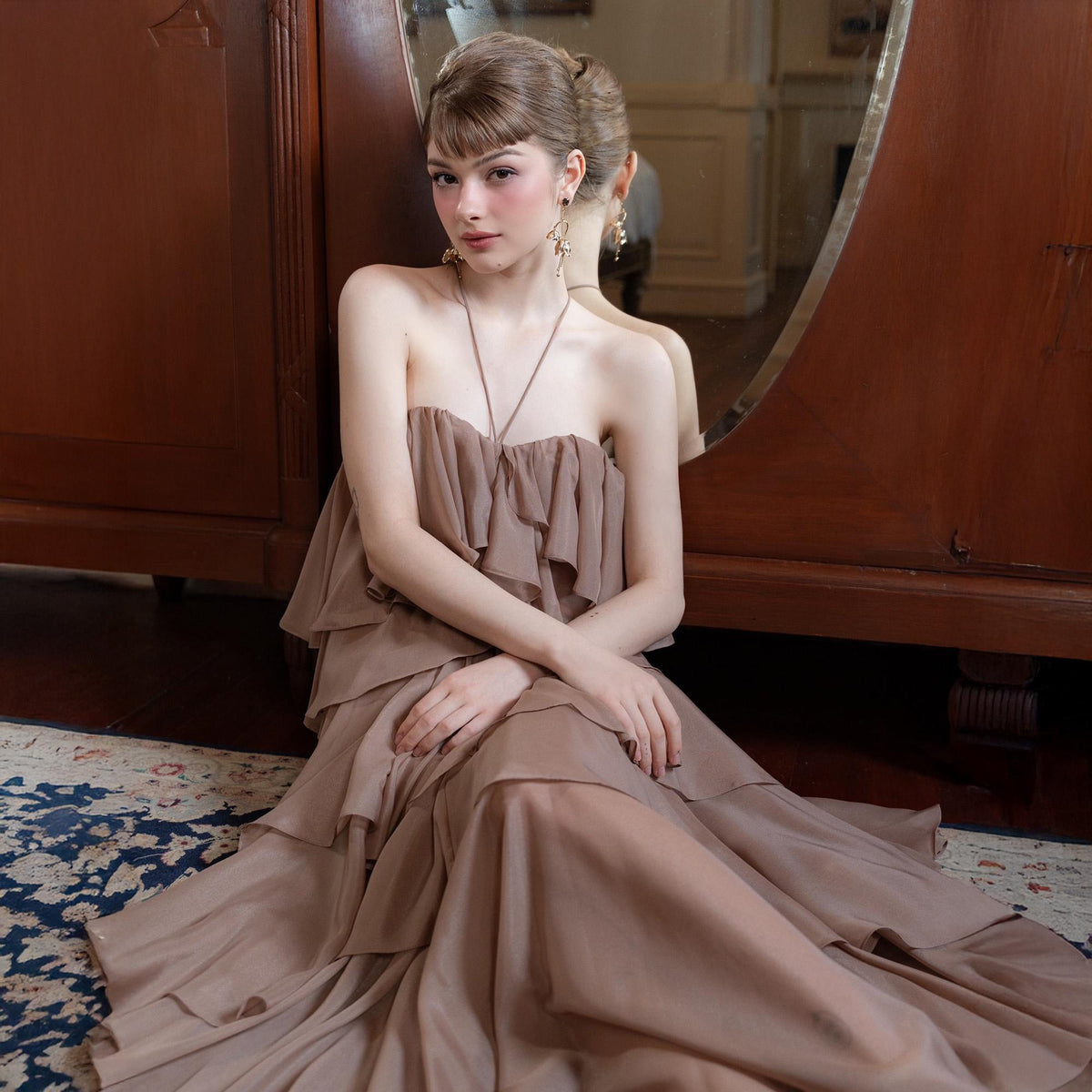 May include: A strapless, layered, taupe-colored dress. The dress features a ruffled bodice and a flowing skirt. The model is sitting on a patterned rug in front of a wooden cabinet and a mirror.