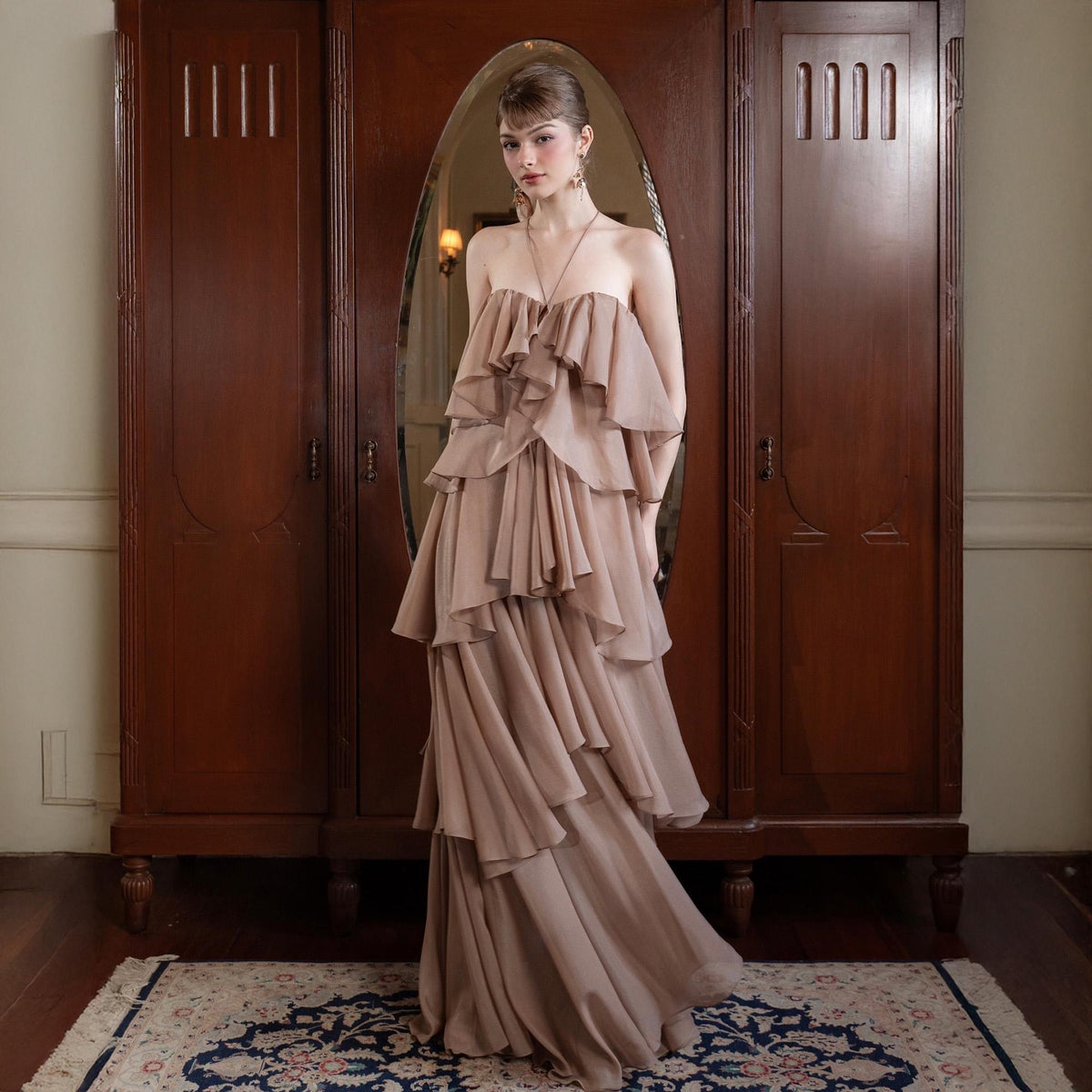May include: A floor-length, strapless gown in a soft, muted rose color. The dress features multiple layers of cascading ruffles, creating a flowing silhouette. The model is standing in front of a dark wood wardrobe with an oval mirror.