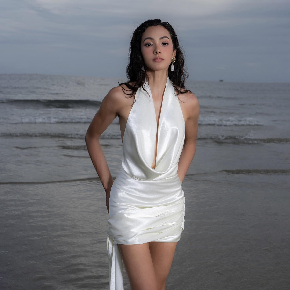 May include: A white satin halter dress with a draped neckline and a gathered skirt. The dress features a low back and a flowing, asymmetrical hemline. The model is standing on a beach with the ocean in the background.