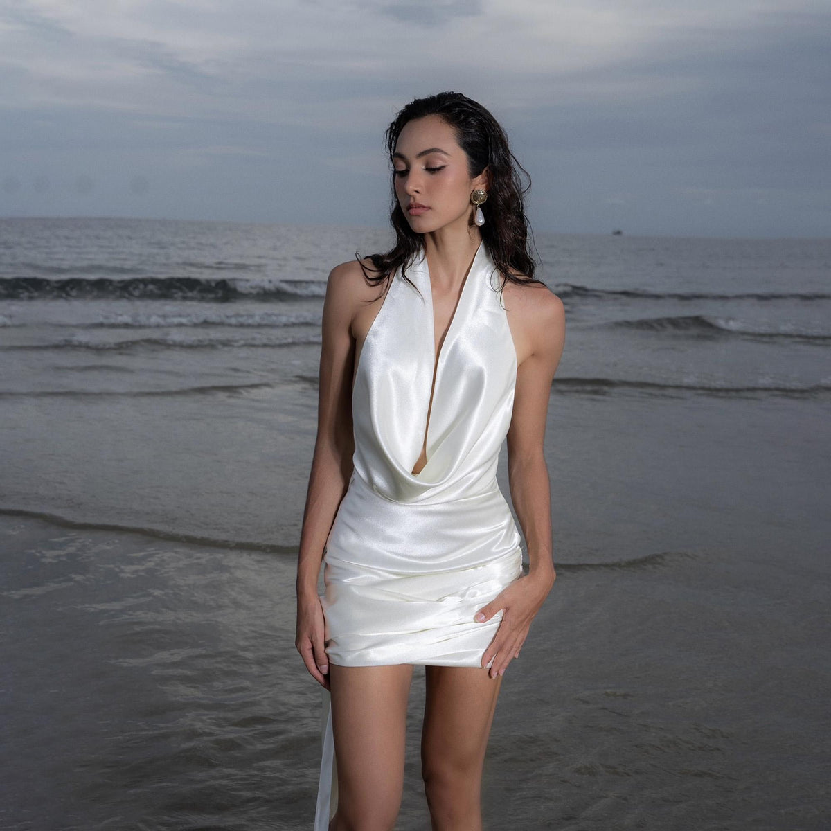 May include: A white satin halter dress with a cowl neckline. The dress is short, with a draped skirt and a flowing train. The model is standing in the ocean with the dress on. The background is a cloudy sky and the ocean.