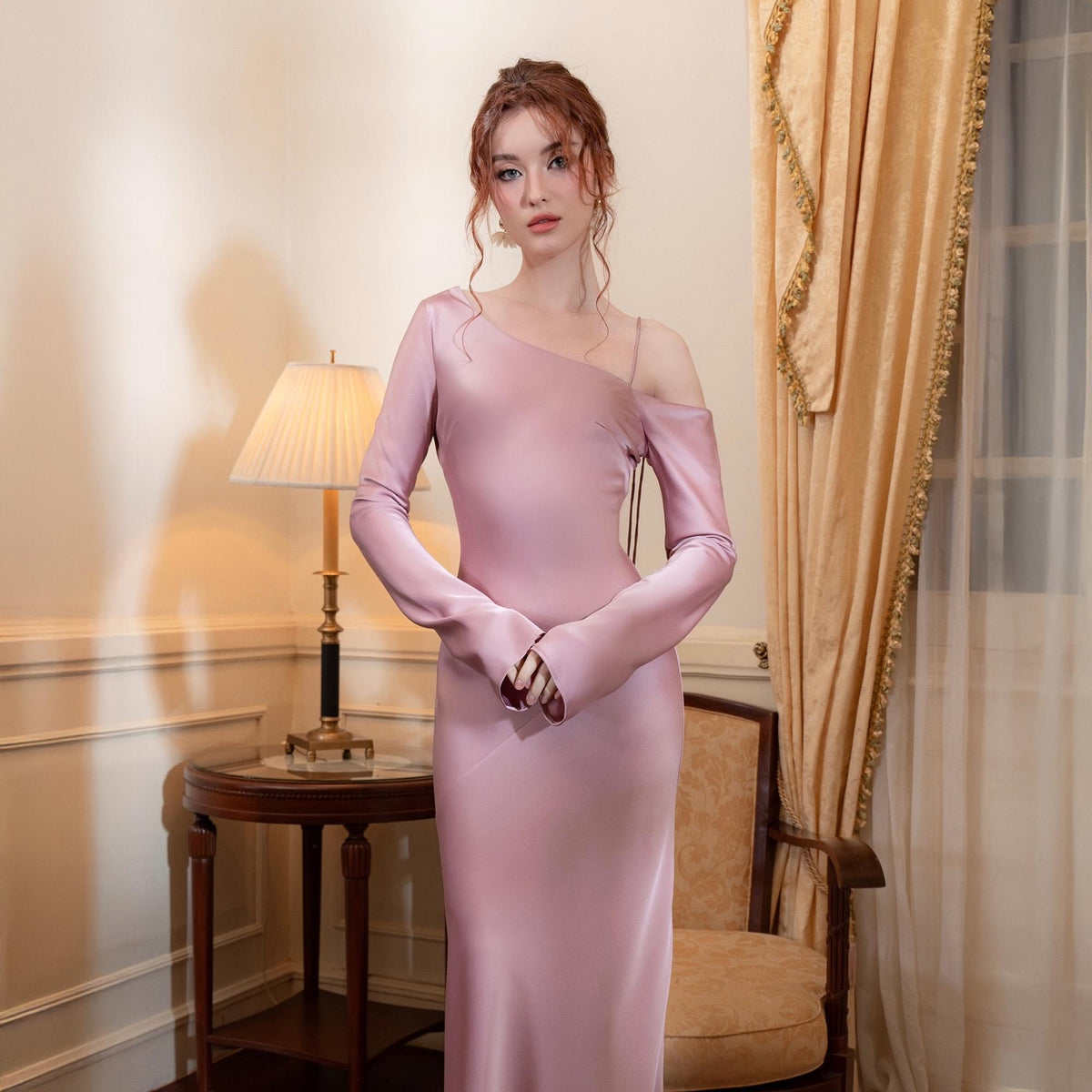 May include: A long, pale pink satin dress with a one-shoulder design and long sleeves. The dress has a fitted bodice and a flowing skirt. The model is standing in a room with a lamp and a chair. The dress is elegant and suitable for formal events.
