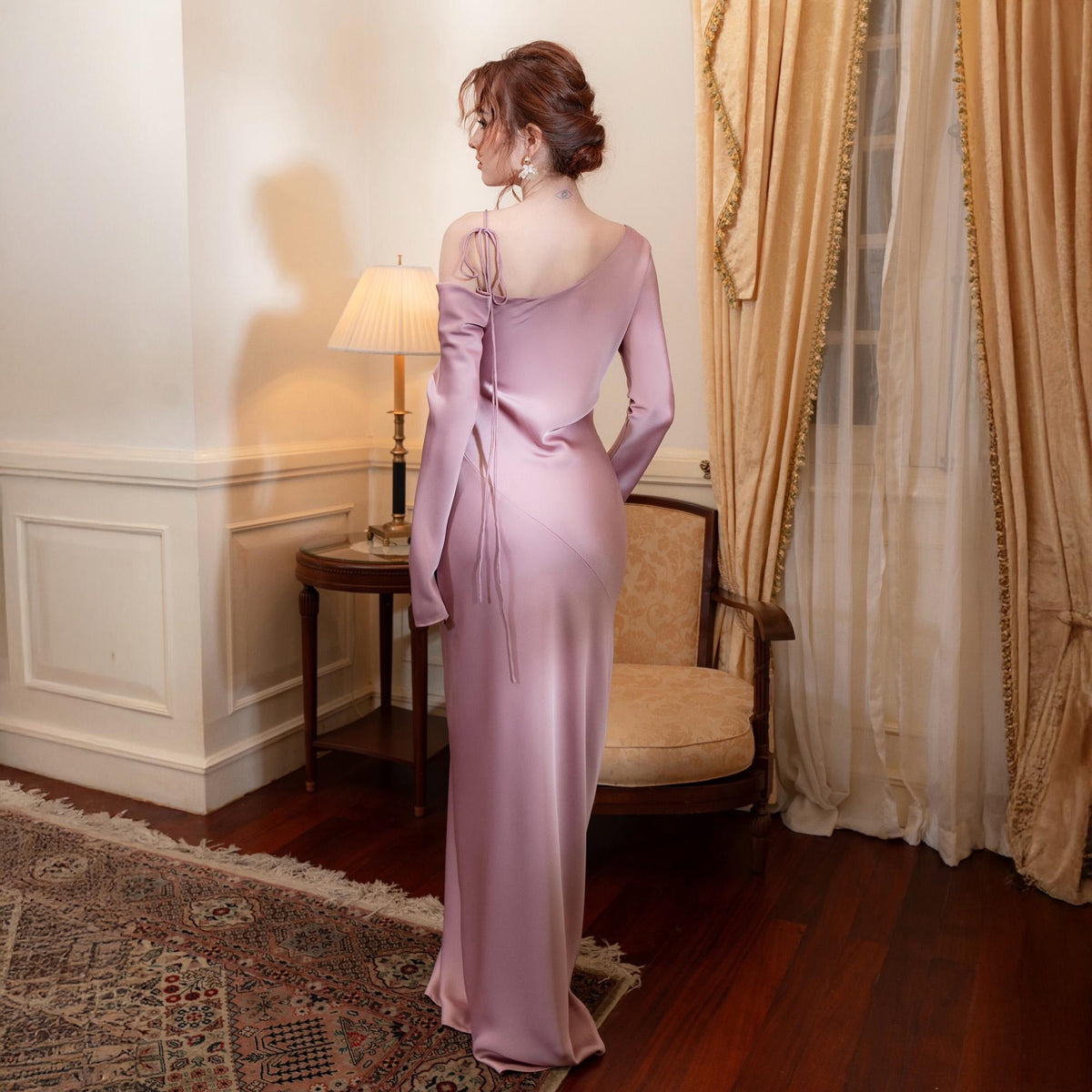 May include: A long, flowing, pale pink satin dress with a one-shoulder design and long sleeves. The dress features a draped neckline and a subtle train. The model is standing in a room with a patterned rug, a side table, and a lamp.