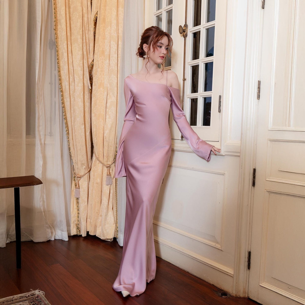 May include: A long, dusty rose-colored satin gown with a one-shoulder neckline and long sleeves. The dress features a fitted bodice that flows into a floor-length skirt. The sleeves have a flared design. The model is standing near a white door and a window with beige curtains.
