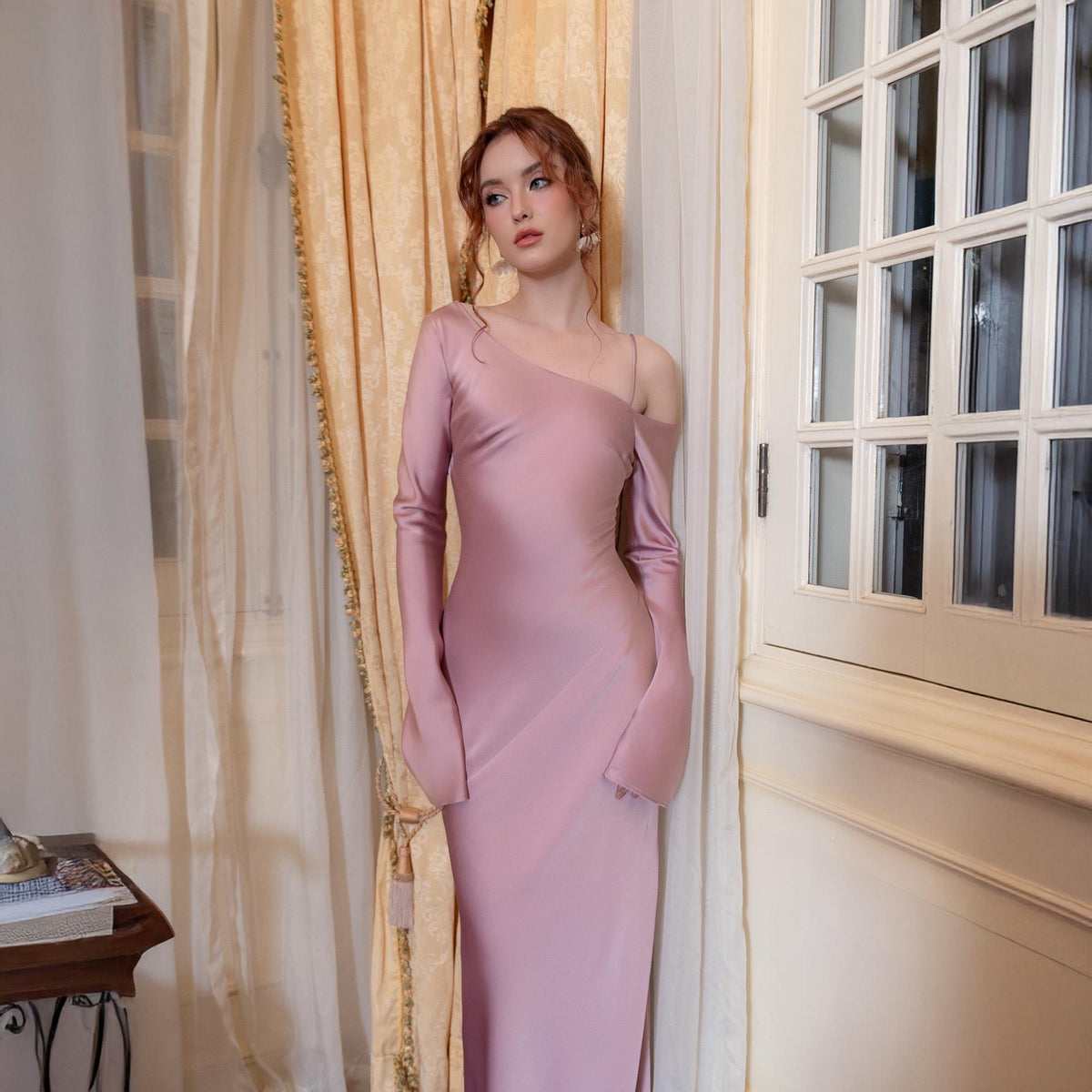 May include: A dusty rose, floor-length gown with an asymmetrical neckline and one long sleeve. The dress features a flowing, bell-shaped sleeve on the right arm and a delicate strap on the left shoulder. The dress is made of a smooth, silky fabric.