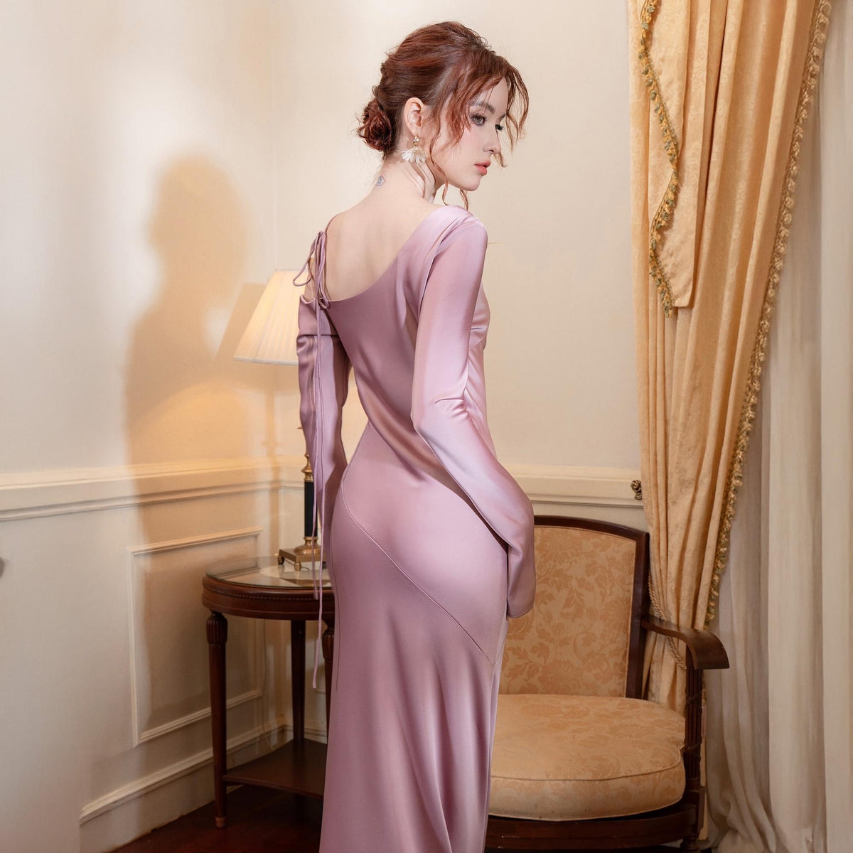 May include: A long, dusty rose-colored satin dress with a one-shoulder design and long sleeves. The dress has a fitted silhouette and a subtle diagonal seam across the front. The model is standing near a table and chair.
