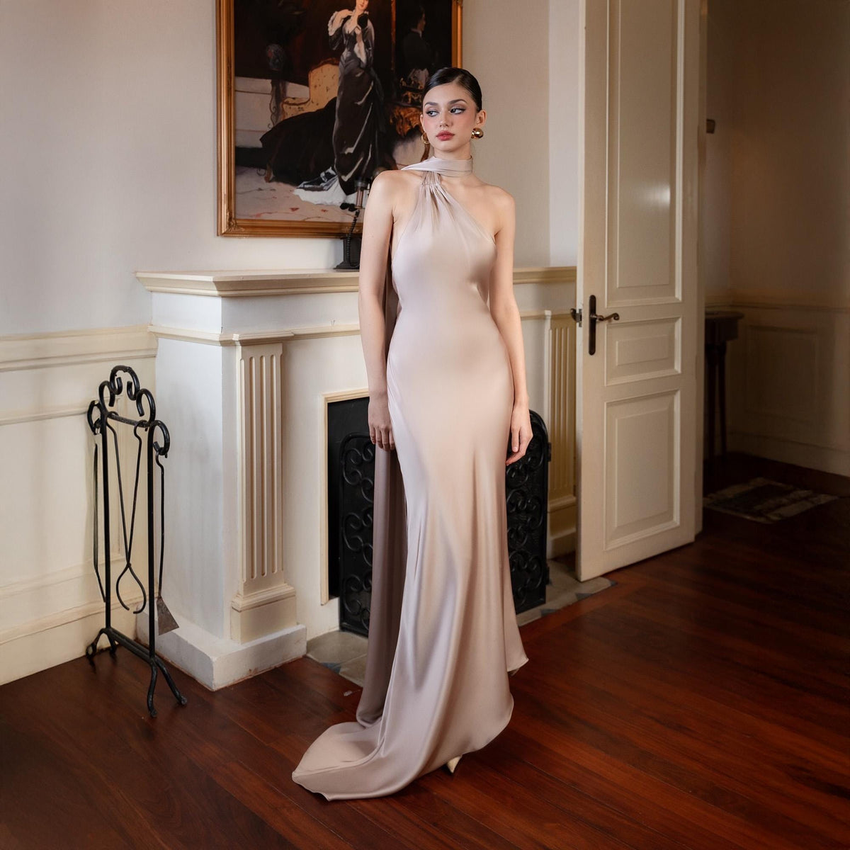 May include: A floor-length, pale pink satin gown with a halter neckline and a flowing train. The dress is form-fitting through the bodice and flares out at the hem. The background includes a fireplace, artwork, and a wooden floor.