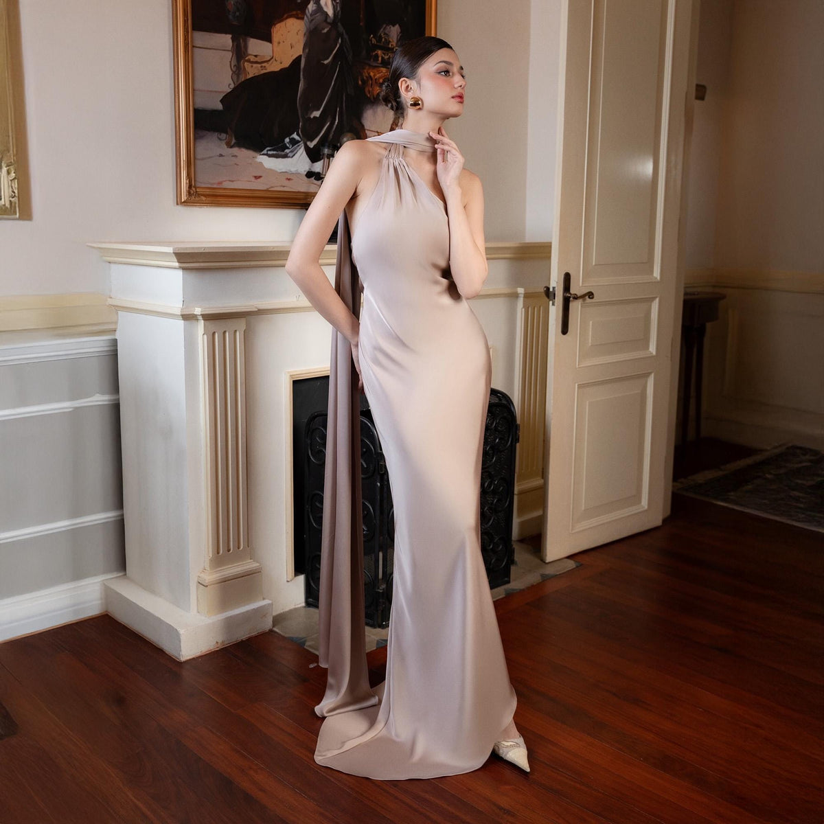 May include: A floor-length, light beige satin gown with a halter neckline and a flowing cape detail. The dress has a mermaid silhouette and is worn with white heels. The background includes a fireplace, artwork, and a wooden floor.