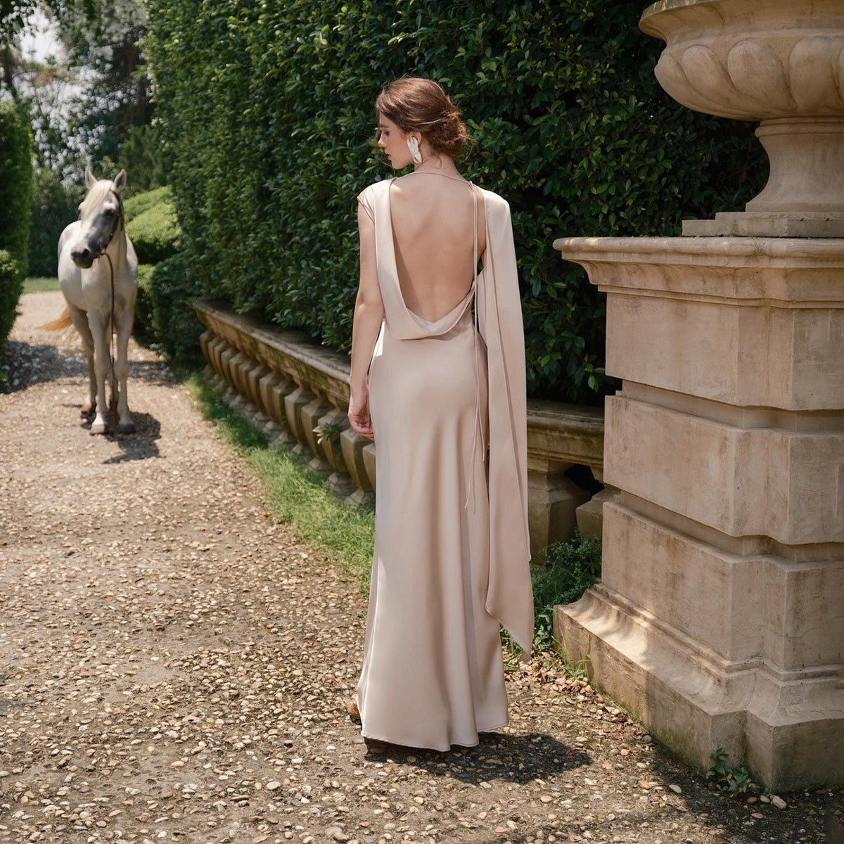 May include: A beige, backless formal gown with a draped shoulder detail. The dress is floor-length and made of a flowing fabric. The image is taken outdoors, with a horse in the background.