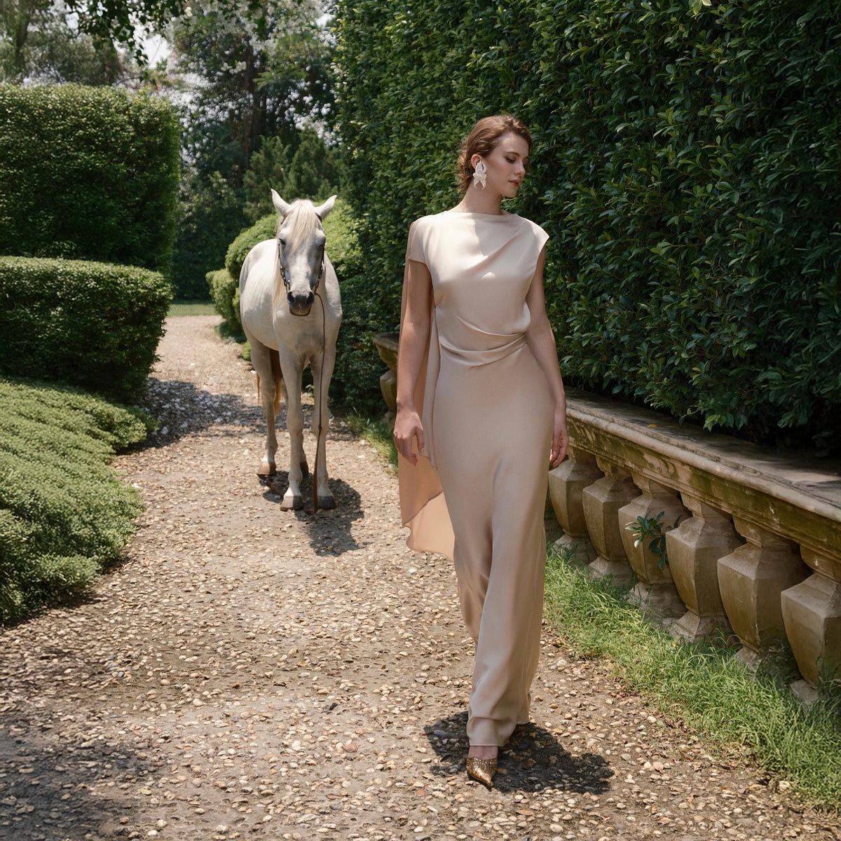 May include: A woman in a beige gown walks on a gravel path, a white horse follows behind. The dress has a draped bodice and a flowing skirt. The scene is set in a garden with green hedges and a stone wall.