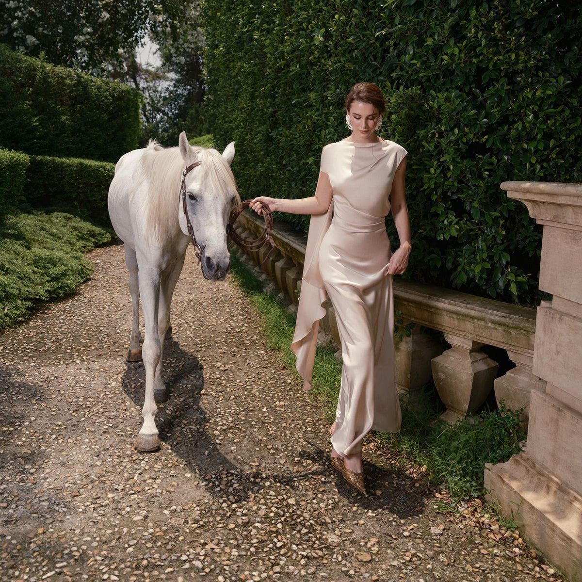 May include: A woman in a flowing, pale peach-colored dress walks alongside a white horse on a gravel path. The dress has a draped neckline and a cape-like detail. The horse wears a bridle. The scene is set outdoors with greenery and stone accents.