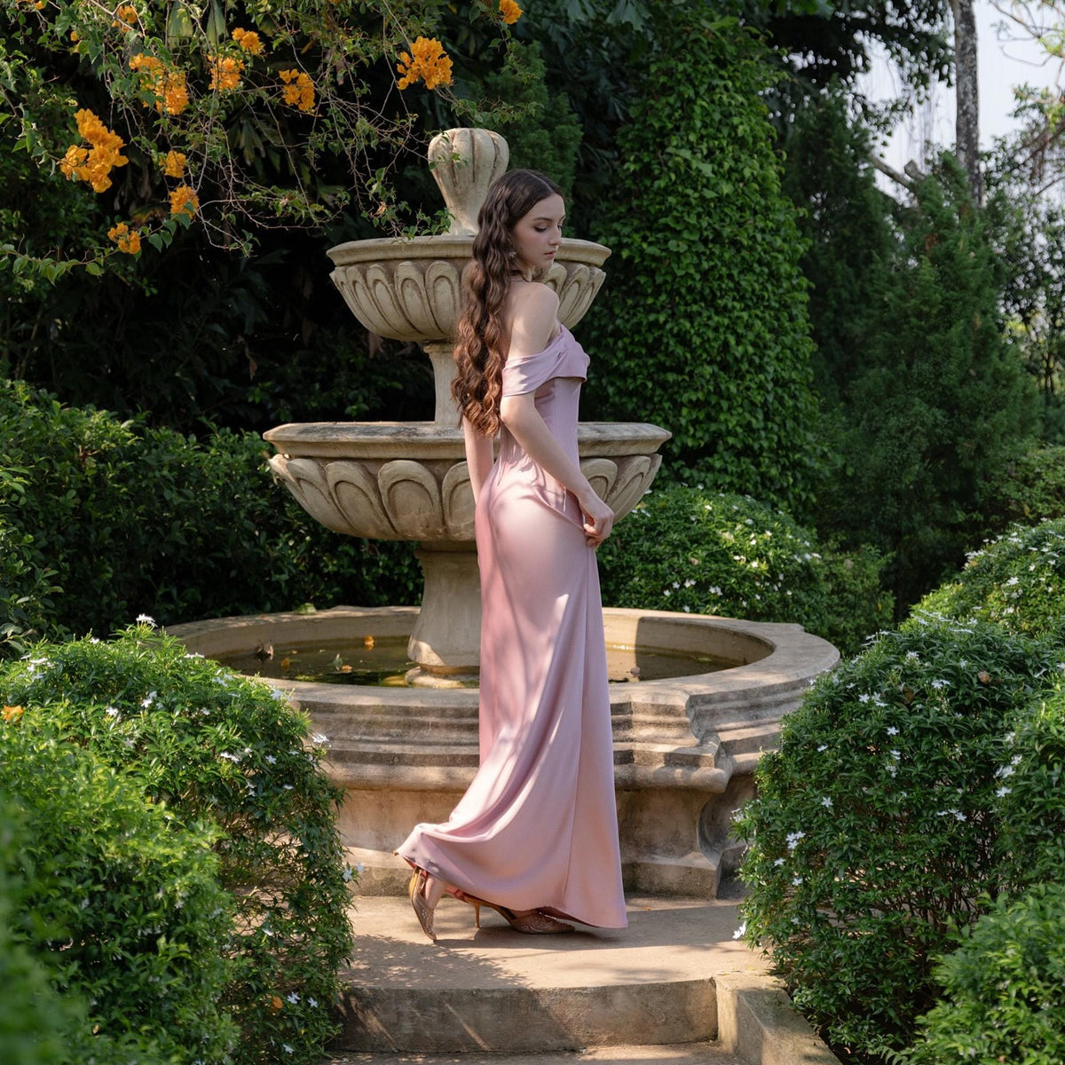 May include: A long, flowing, pale pink dress with off-the-shoulder sleeves. The dress is worn outdoors near a tiered stone fountain surrounded by lush greenery and yellow flowers. The dress is floor-length and appears to be made of a silky material.