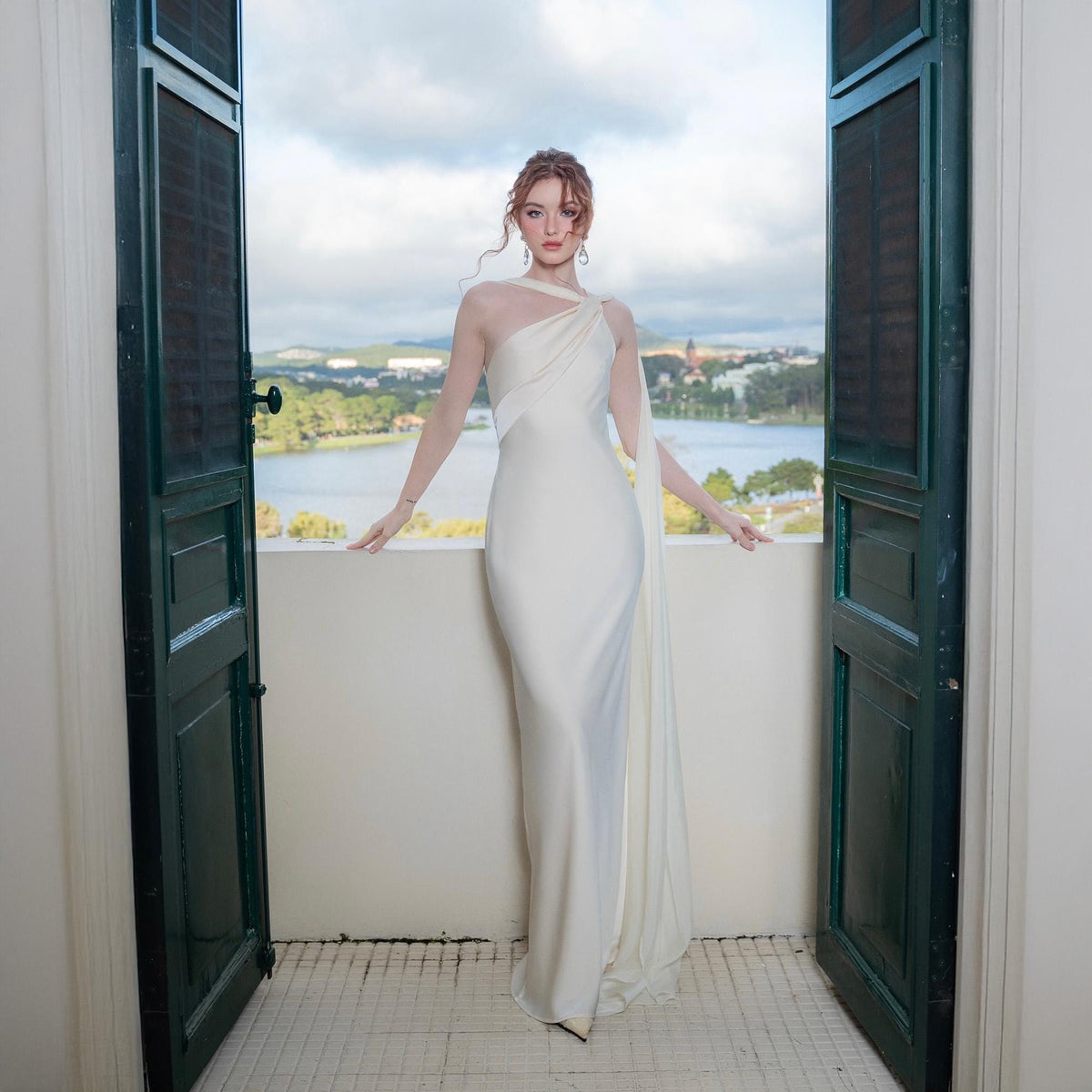 May include: A white, floor-length gown with a one-shoulder design and a flowing train. The dress features a sheer neckline and sleeves. The image is framed by open, dark green doors, with a scenic lake and town visible in the background.