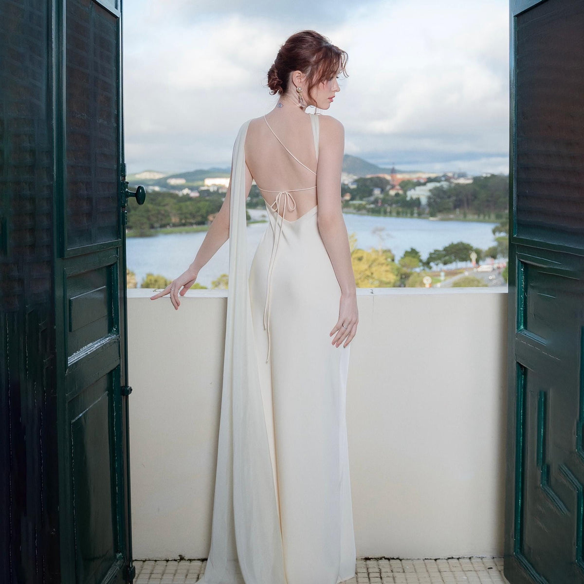May include: A cream-colored, floor-length gown with a draped shoulder and open back. The dress features a flowing cape detail and delicate tie closure. The woman is standing on a balcony overlooking a lake and mountains.